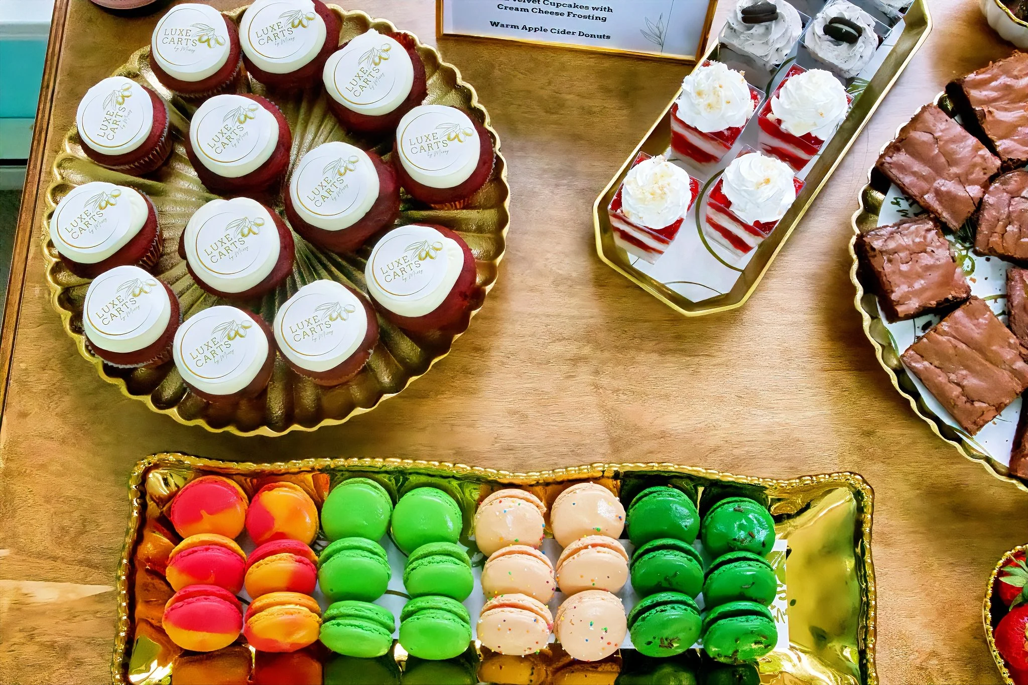 Beautiful desserts displayed on gold trays, including red velvet cupcakes & personalized logos and colorful macarons in pink, yellow, white with sprinkles, and green flavors on dessert cart at a wedding
