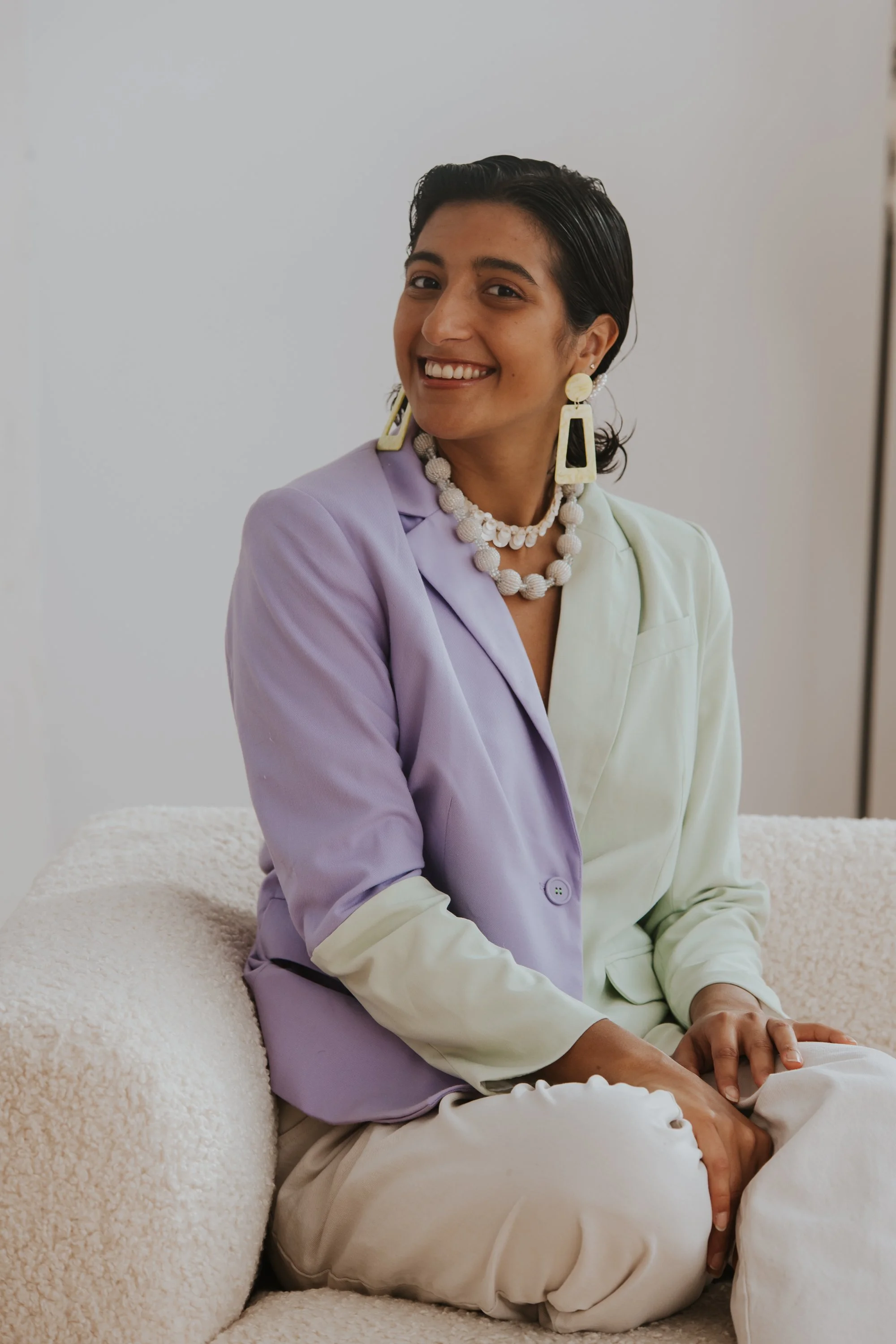Woman smiling at the camera. She has dark hair and is wearing large earrings, a chunky pearl necklace, and a purple and light green blazer