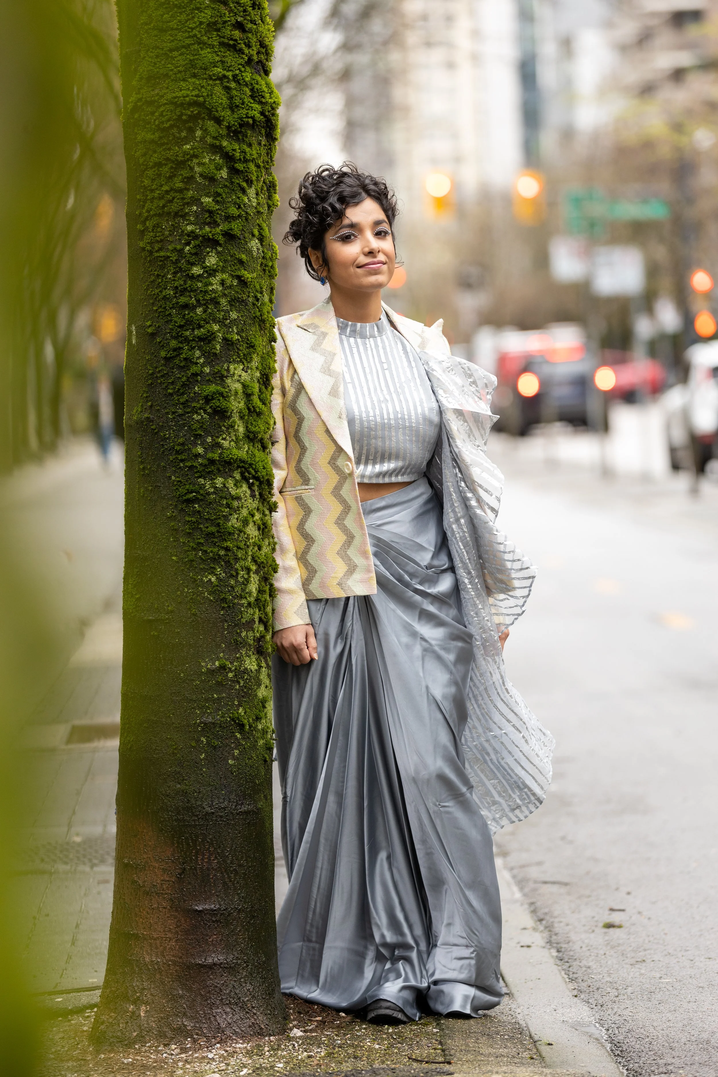 Woman staring at the camera while leaning on a tree. She is wearing a zig zag stripe jacket, a blue and white vertical lines shirt, and a grey skirt