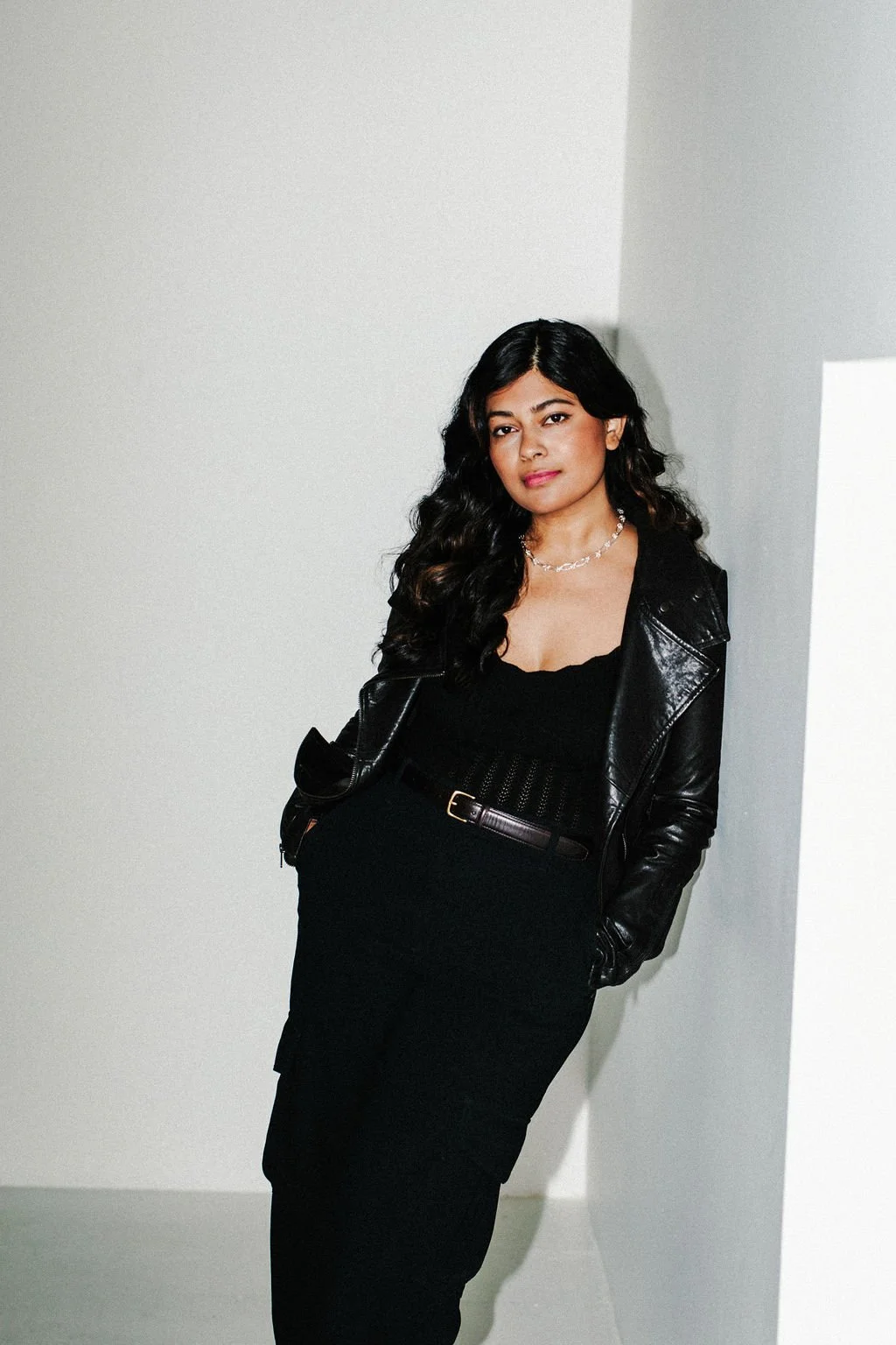 Woman leaning against a white wall. She has her hands in her pockets. She has black hair and is wearing a black jacket, black shirt, and black skirt