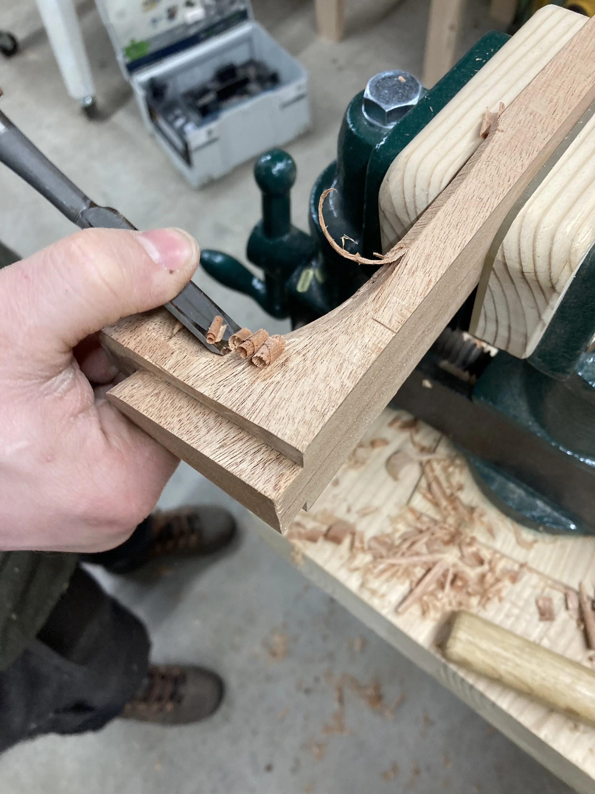 Person using a chisel on a wooden board secured in a vise, producing wood shavings.