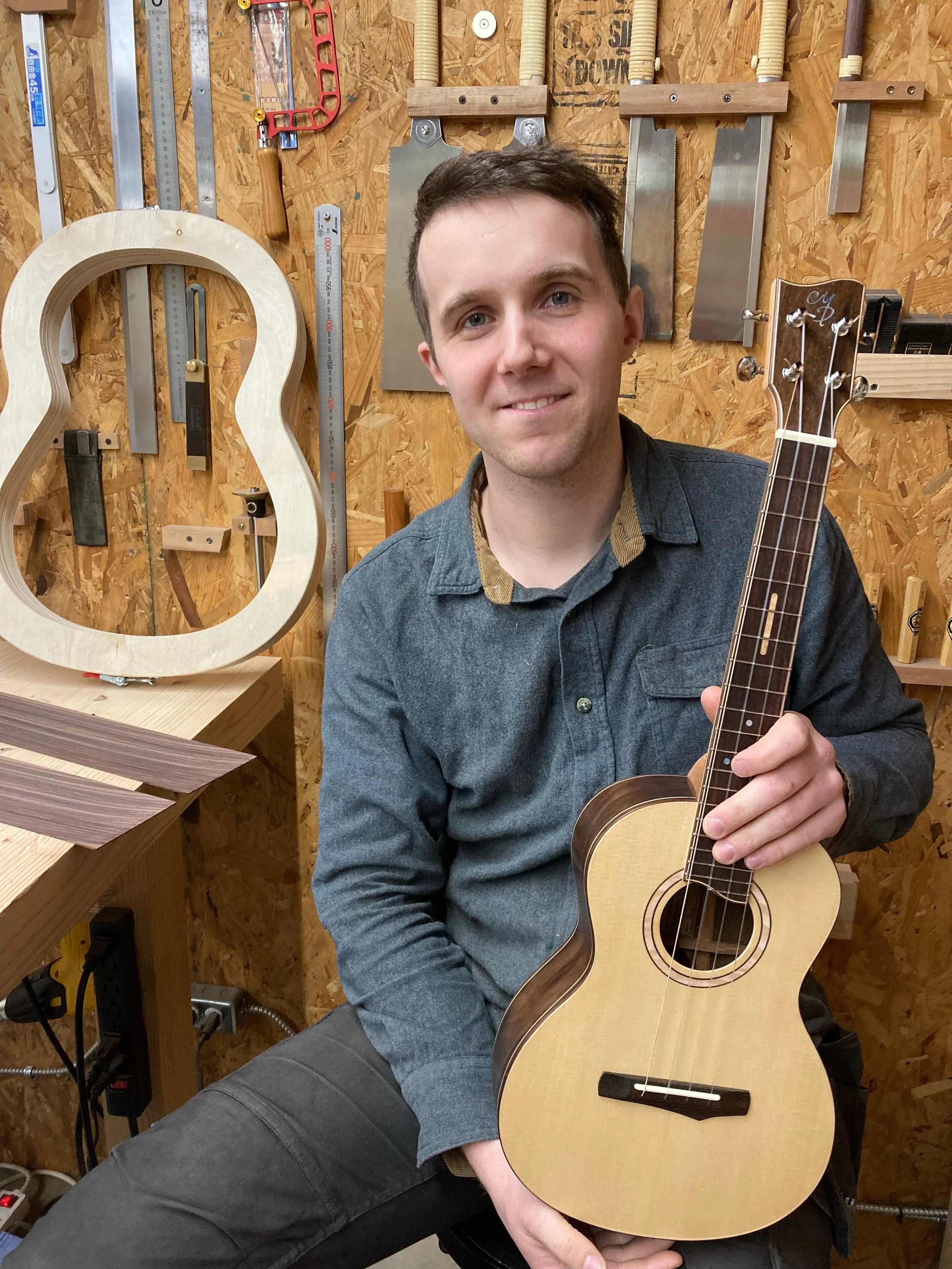 Person holding a ukulele in a woodworking shop, surrounded by tools and guitar frame on the wall.