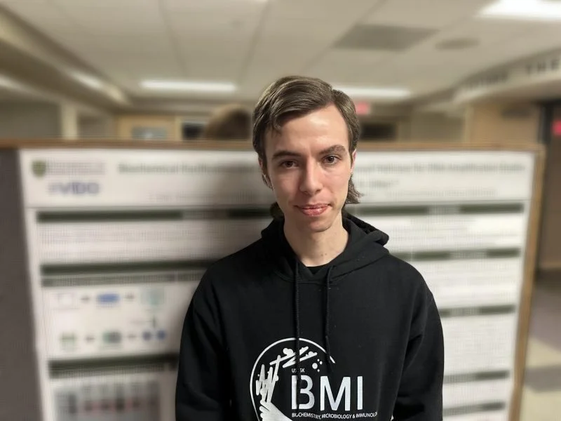 Young man with brown hair wearing a black hoodie with 'BIM' logo, standing in front of a scientific poster display at a conference or academic event.