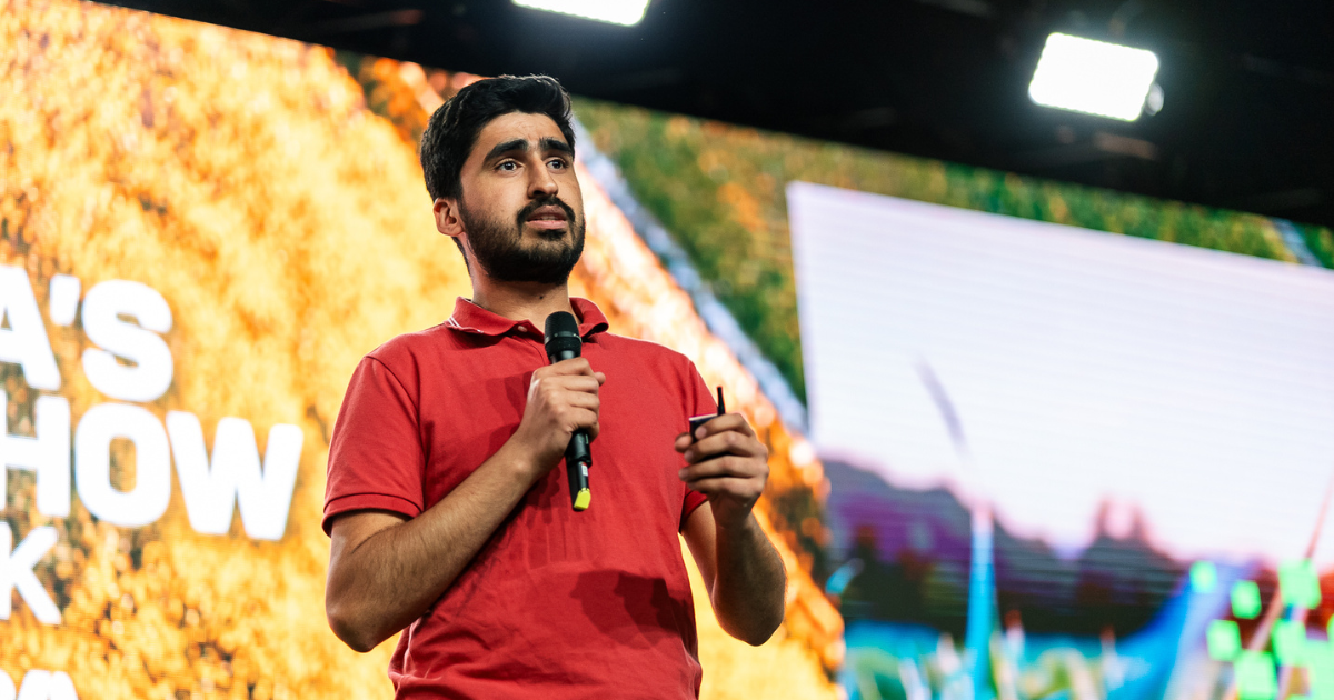A man with dark hair and a beard wearing a red polo shirt, speaking into a microphone on a stage with a large colorful screen in the background.