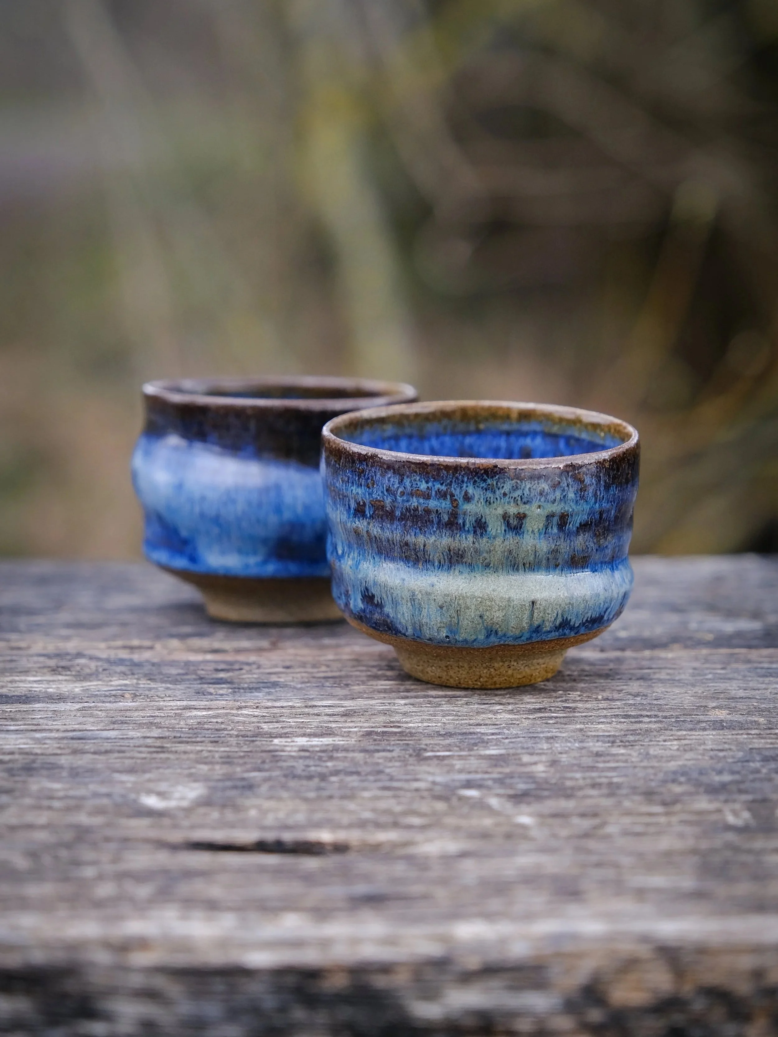 A pair of cups featuring the organic swirly form and flowing blue glaze in a natural setting.