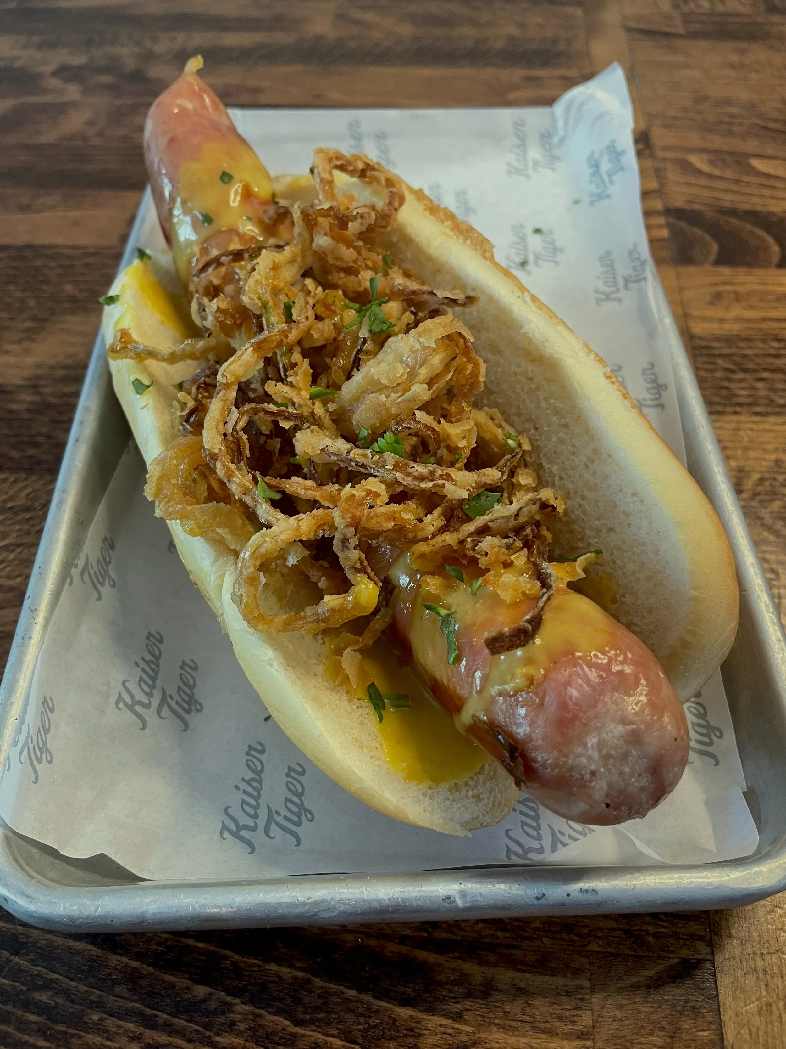 Hot dog with mustard, crispy fried onions, and chives in a bun, served on a tray with branded paper.
