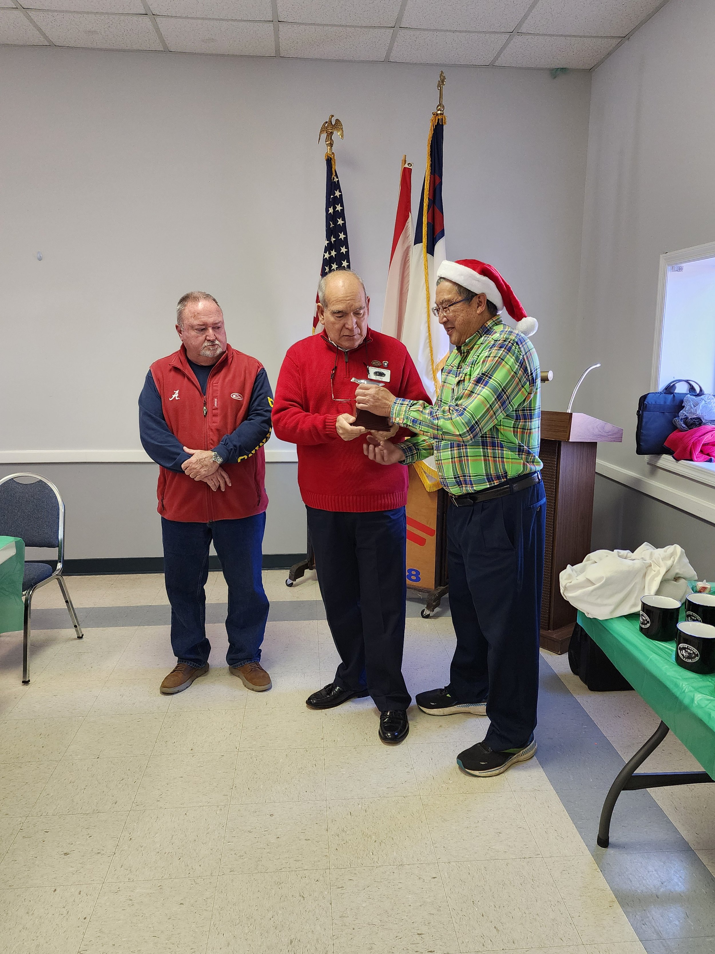 Three men standing indoors, two wearing red sweaters, one with a Santa hat, holding a plaque, with American flags in the background.