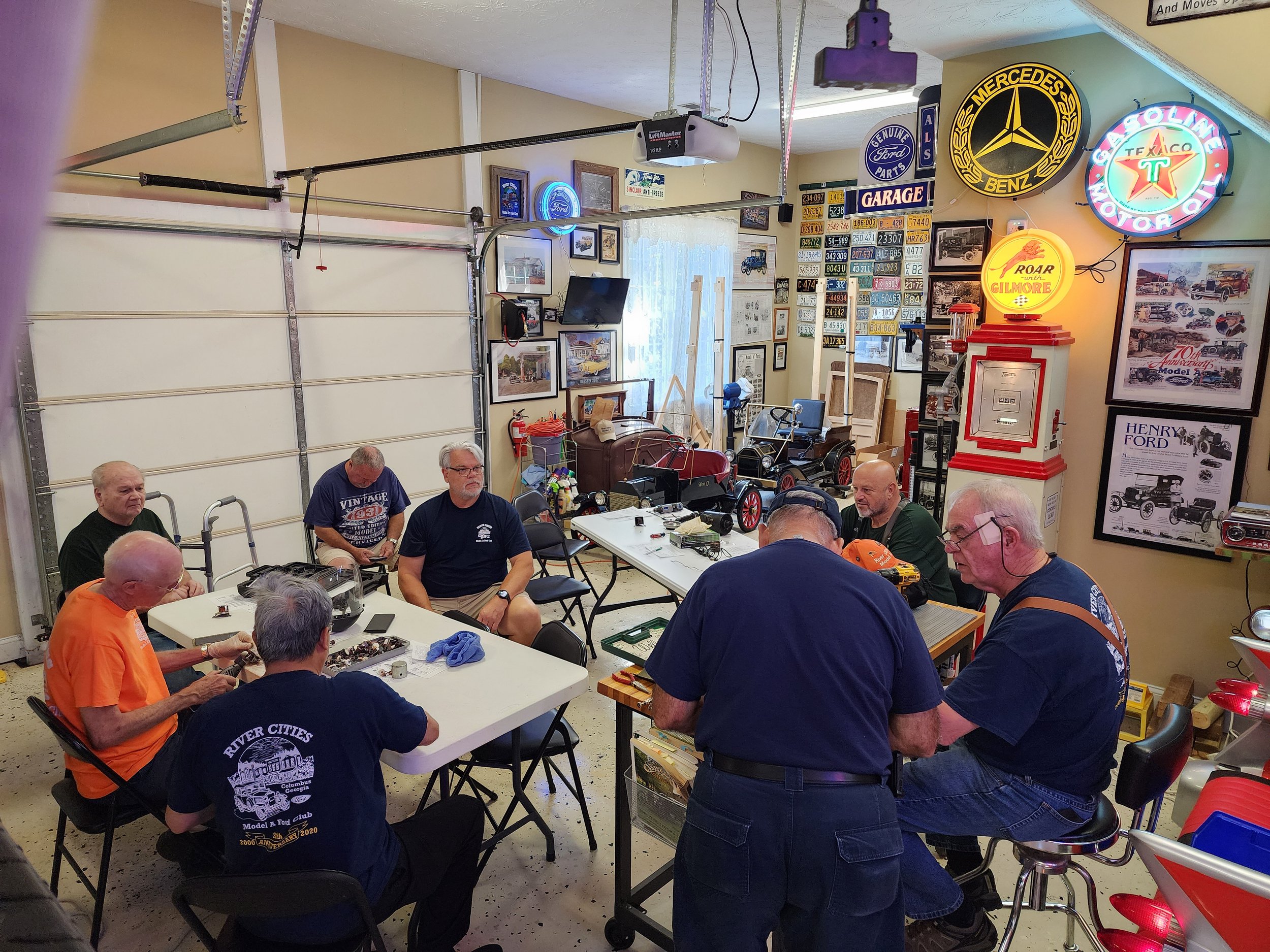 Group of older men gathered in a vintage car-themed garage, sitting around tables with car memorabilia and miniature car models on display. Walls adorned with vintage signs, posters, and license plates. Neon lights and retro decor accent the space.