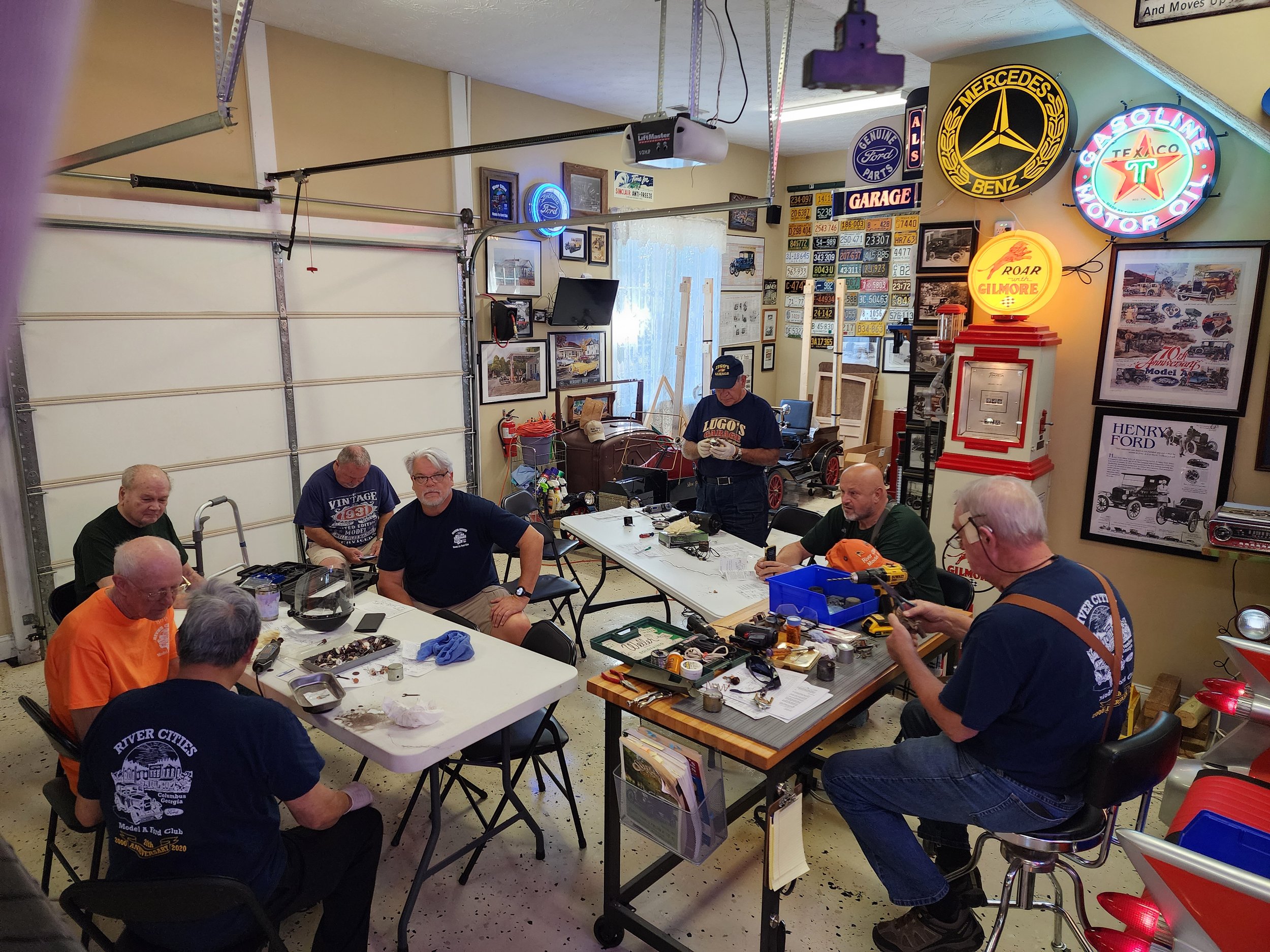 A group of men work on small mechanical projects in a garage filled with vintage automotive memorabilia and signs.