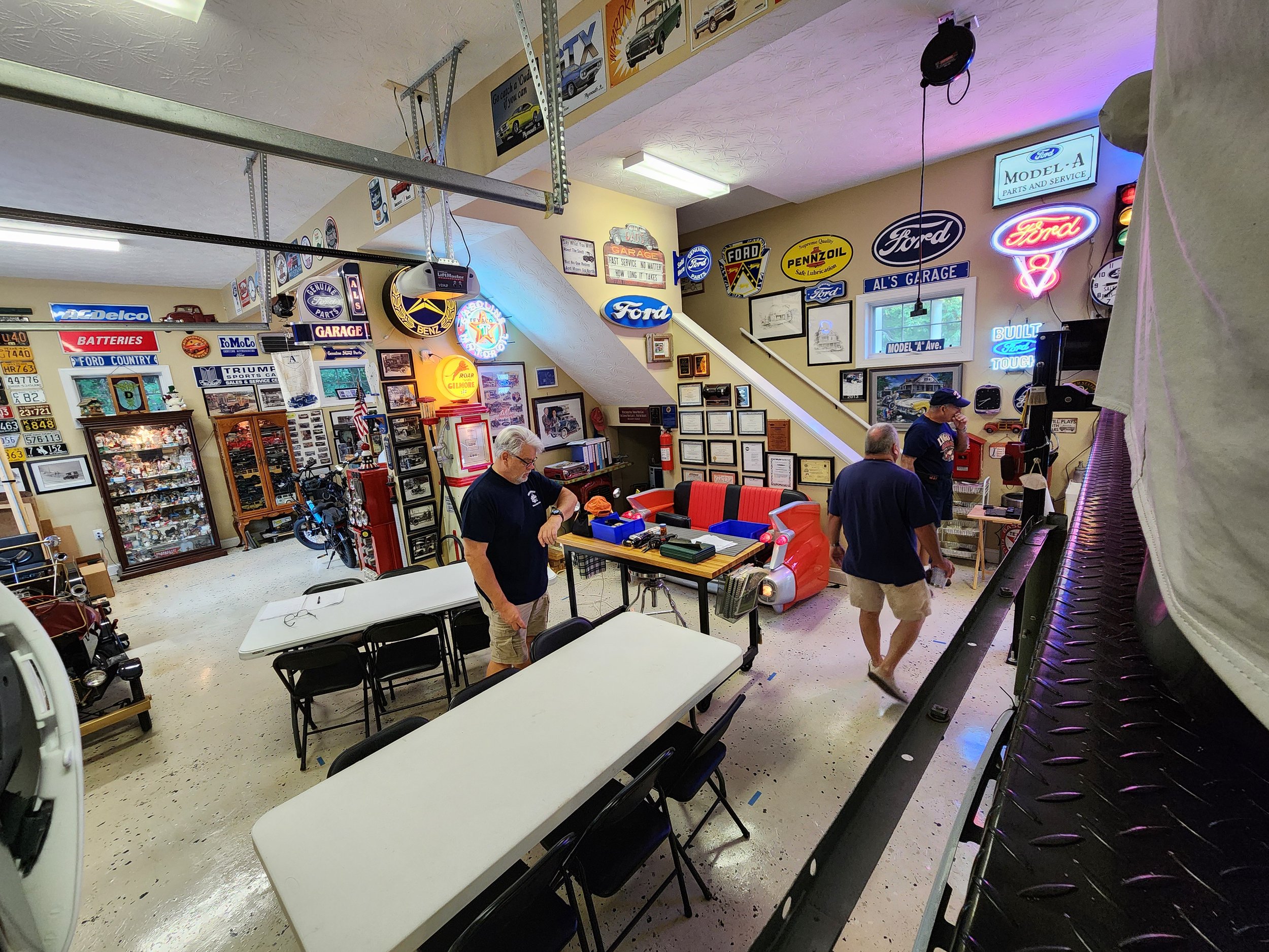 A garage or workshop with vintage automotive signs on the walls, including brands like Ford and Mercedes. There are tables and chairs set up, display cabinets filled with collectibles, and three people in casual attire are present. The room has a cla