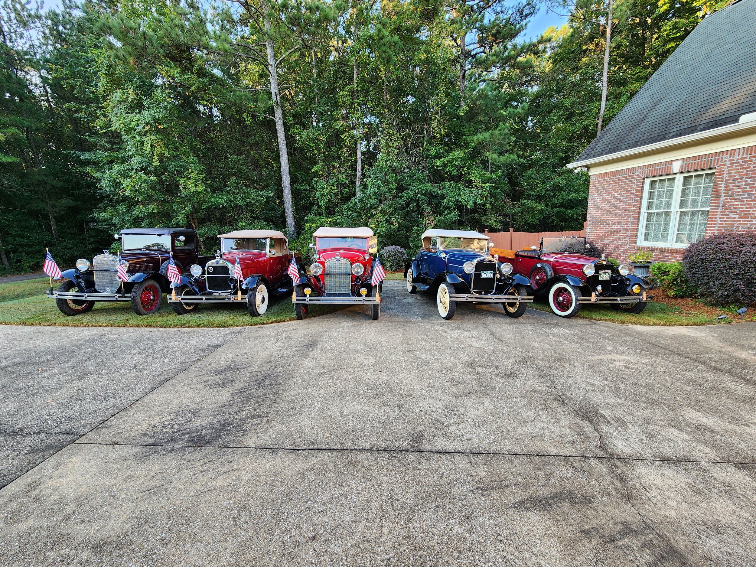 Four vintage cars parked in front of a brick house, adorned with American flags, surrounded by trees and greenery.
