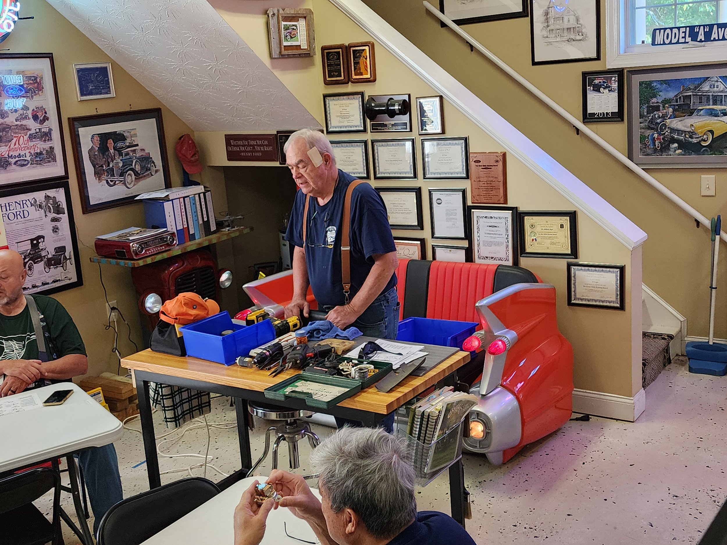 An older man standing by a workbench filled with tools and parts in a room with automotive memorabilia and awards on the walls. Nearby, another person is seated working on a small object. The room features a makeshift automotive-themed seating area a