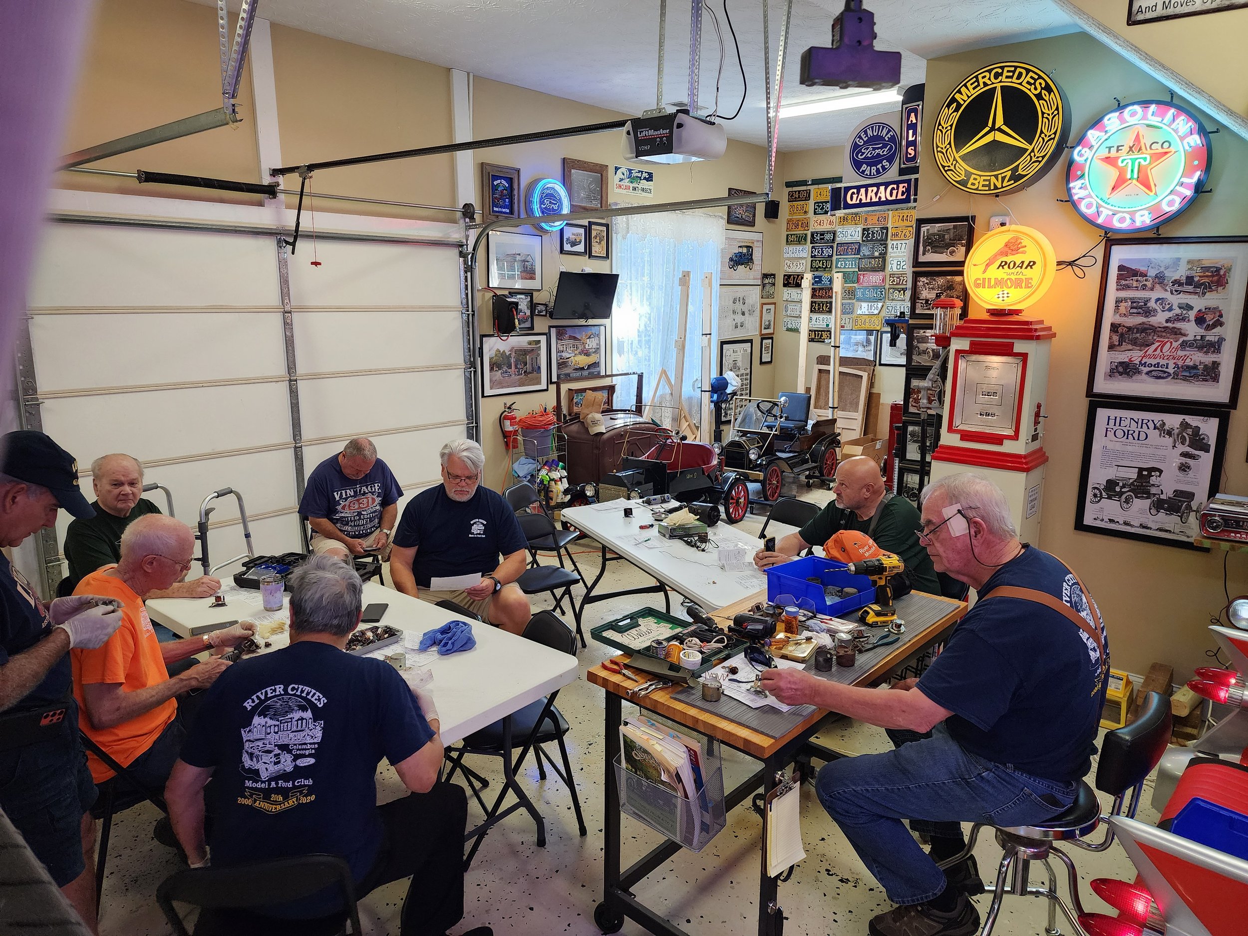Group of men working on model cars in a garage with automotive memorabilia and signs on the walls.