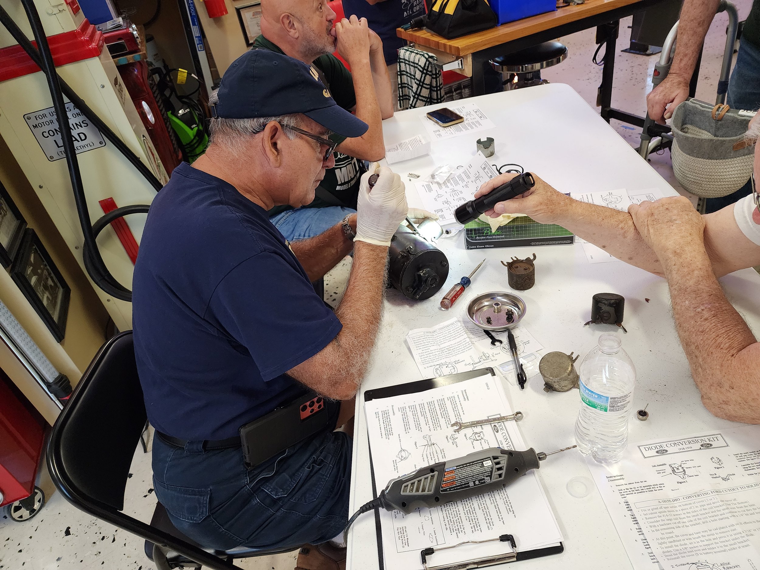 A group of people working on mechanical parts at a table, examining and assembling components with tools and papers. A person is holding a flashlight, and various tools and instruction sheets are spread out on the table. A water bottle and a drill ar
