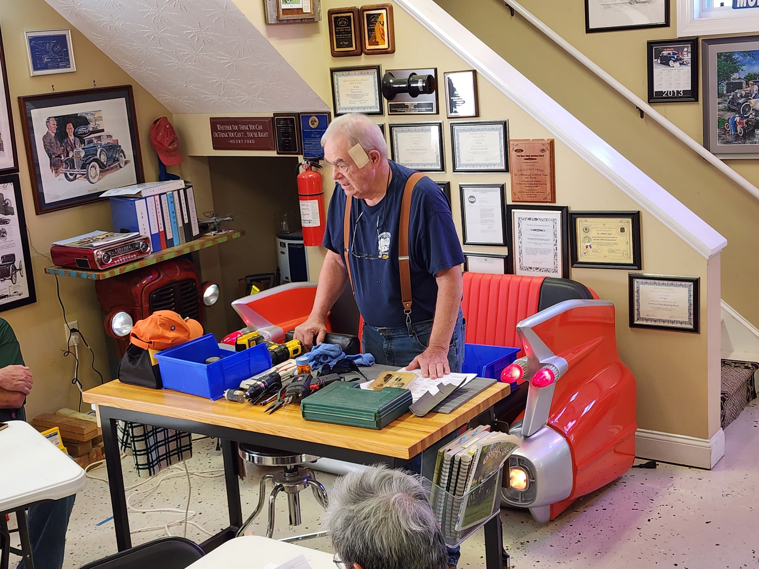Older man standing at a workbench with tools in a room decorated with automotive memorabilia and vintage car parts, including a red car-themed sofa.