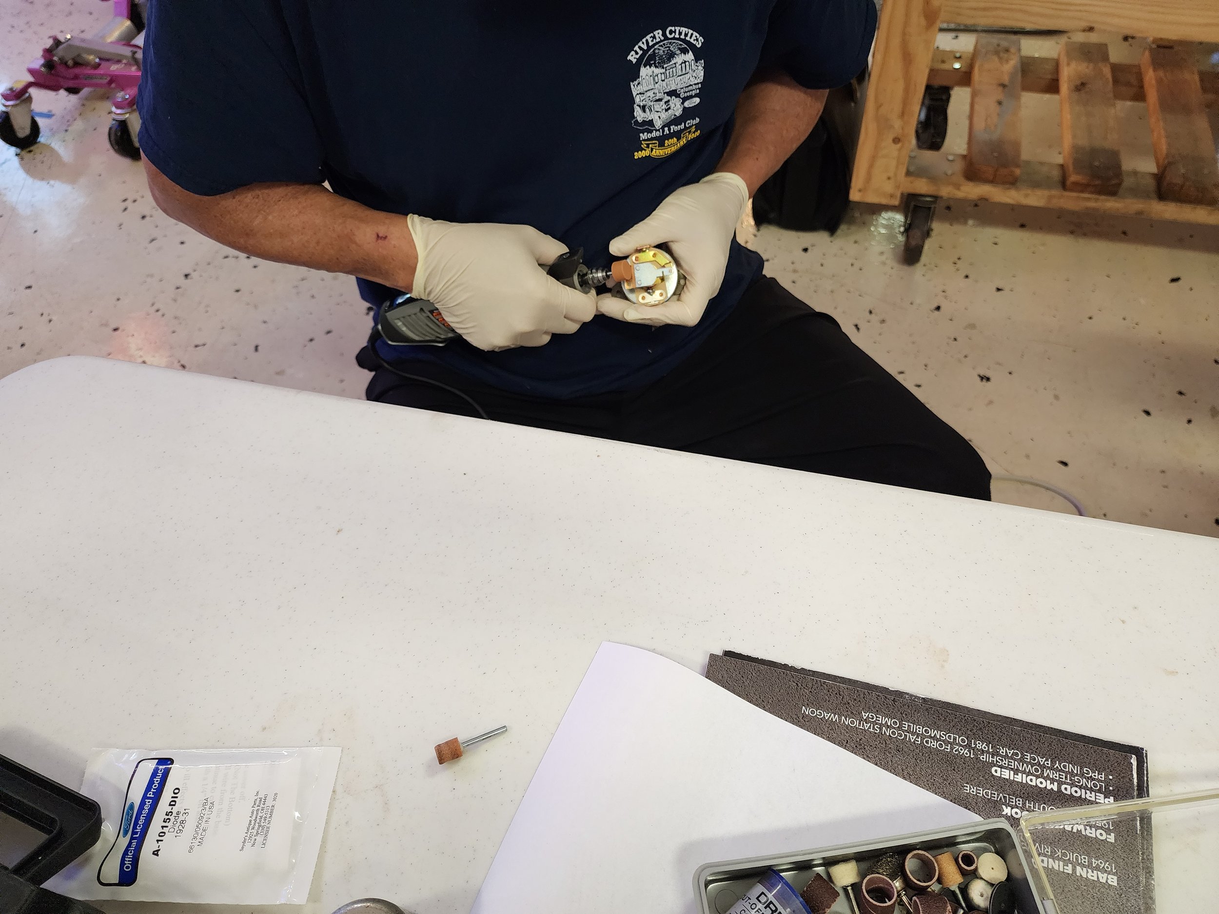 Person wearing gloves using a handheld rotary tool to work on a small object at a table.
