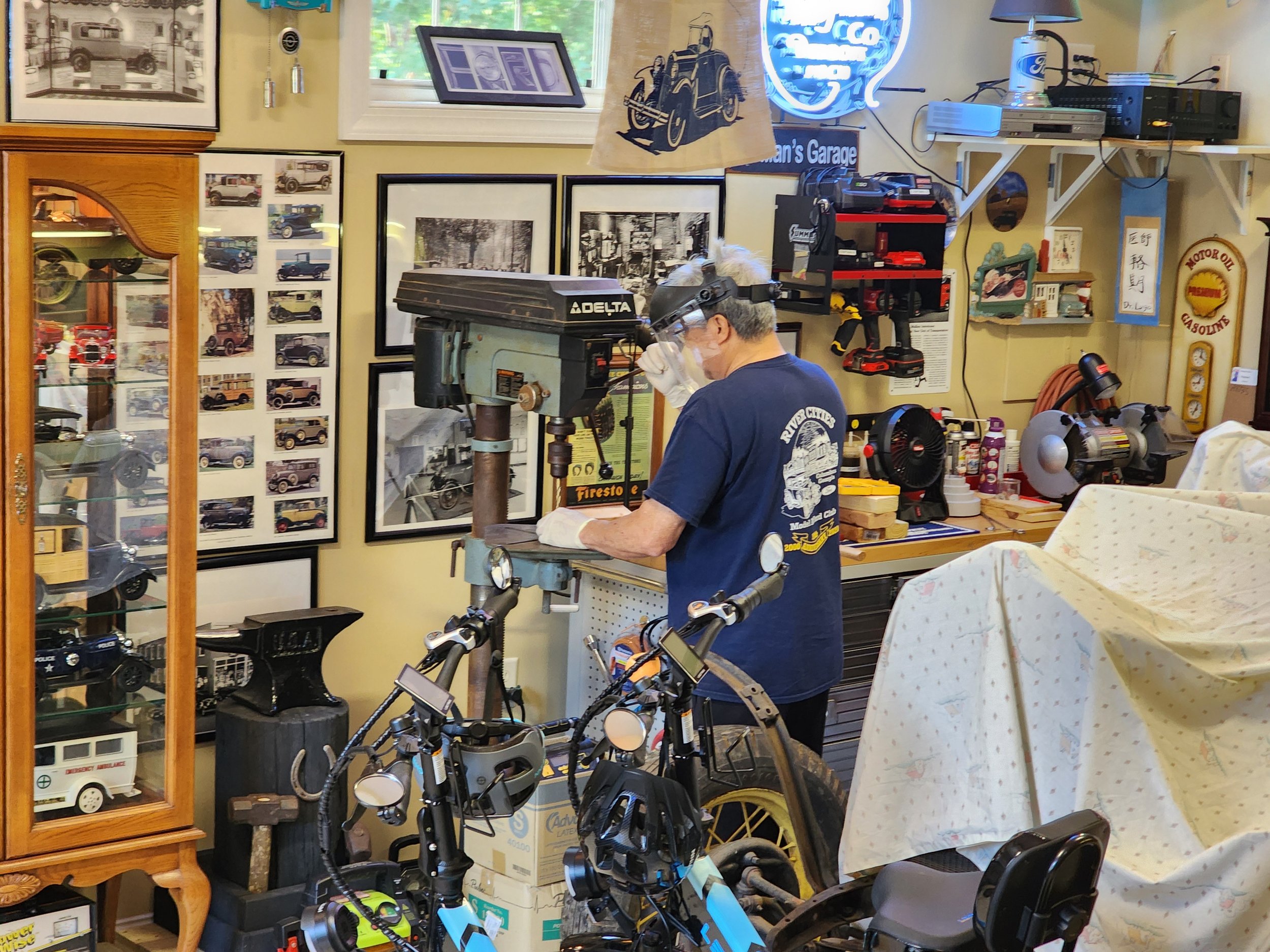 A man working in a garage workshop filled with vintage car memorabilia and tools. He is operating a Delta drill press machine. Walls are decorated with framed automotive photographs and collectibles. Several bicycles and covered items are also presen