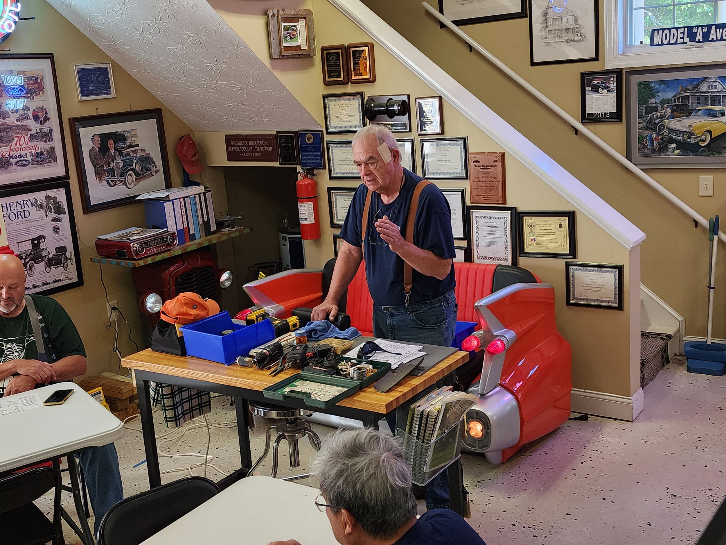 A man giving a presentation in a vintage automobile-themed room with tools on a table in front of him. The walls are decorated with framed certificates and car-related memorabilia. A unique red seat resembling a car's rear end is visible. People are