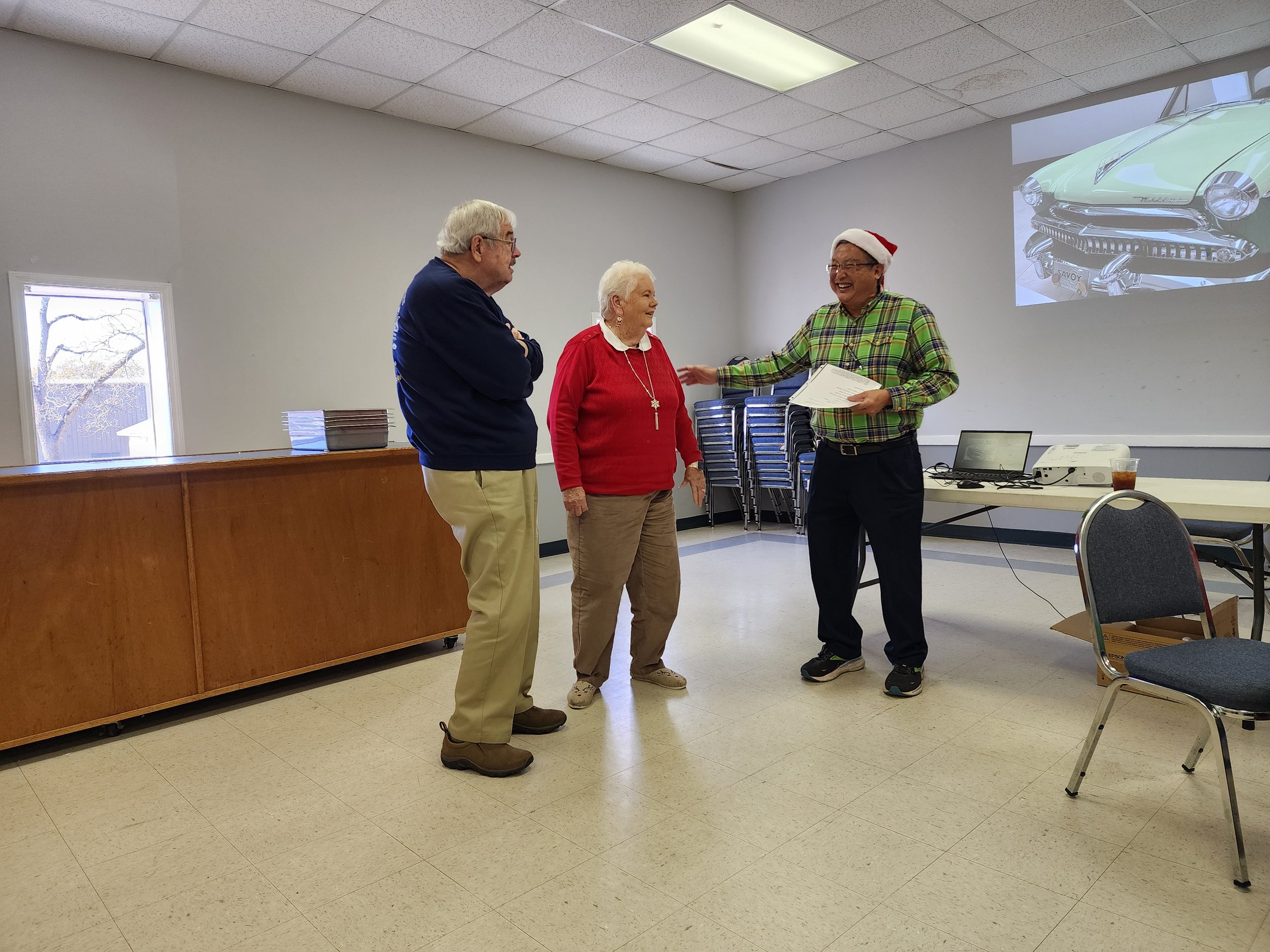 Three people in a room, one wearing a Santa hat holding papers, standing near a table with a laptop and projector. Chairs are stacked in the background and a vintage car image is projected on the wall.