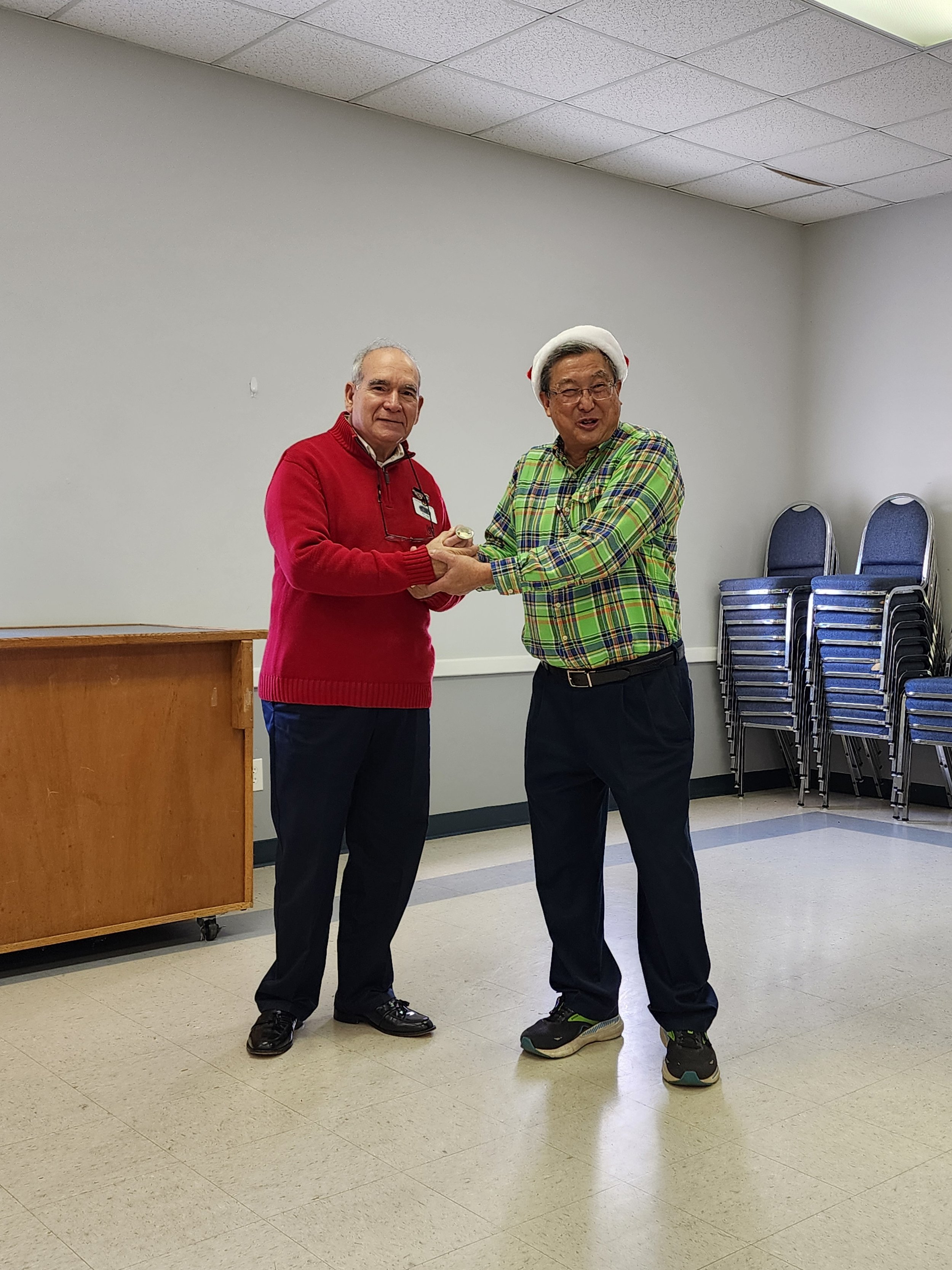Two men in a room; one wearing a red sweater, the other in a festive hat and plaid shirt. They appear to be shaking hands with stacked chairs in the background.