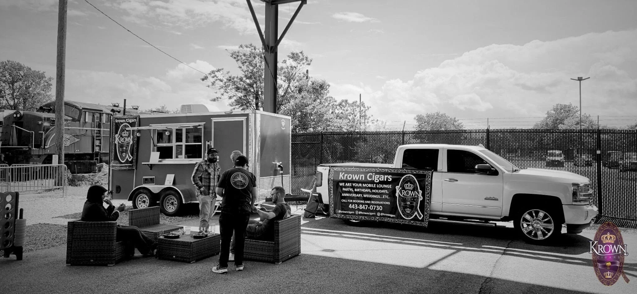 A group of people standing and sitting near a white pickup truck with a banner advertising Krown Cigars, with a food stand, a train, and a fence in the background on a sunny day.
