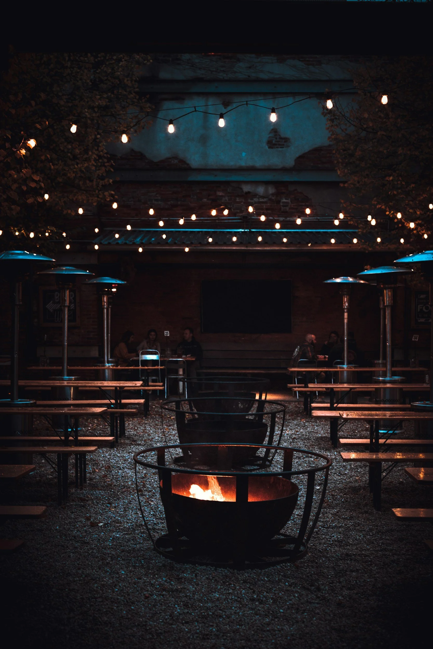 Nighttime outdoor patio with string lights hanging overhead, patio heaters, tables and chairs, a fire pit in the foreground, and a brick wall with a mural of a sky and clouds in the background.