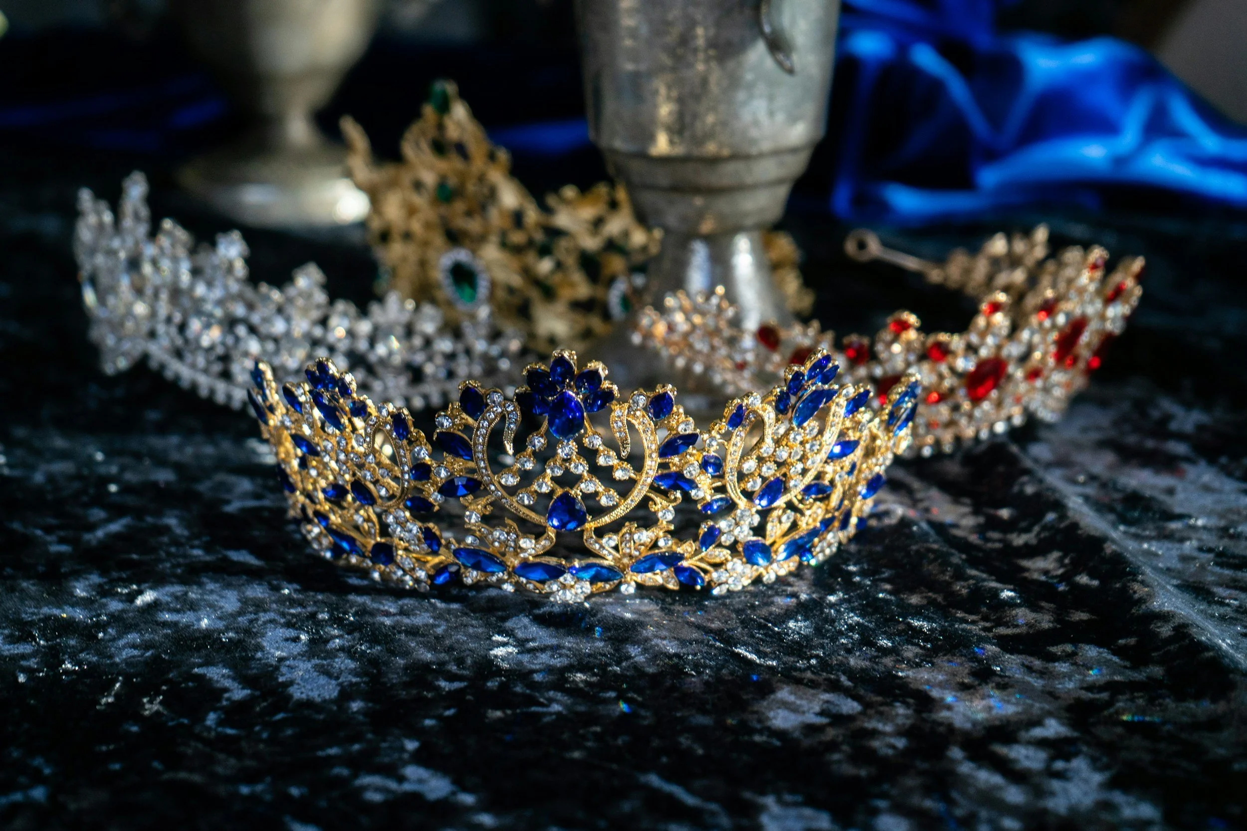 A close-up of a collection of ornate jewelry including tiaras and rings, with a silver container and dark fabric in the background.