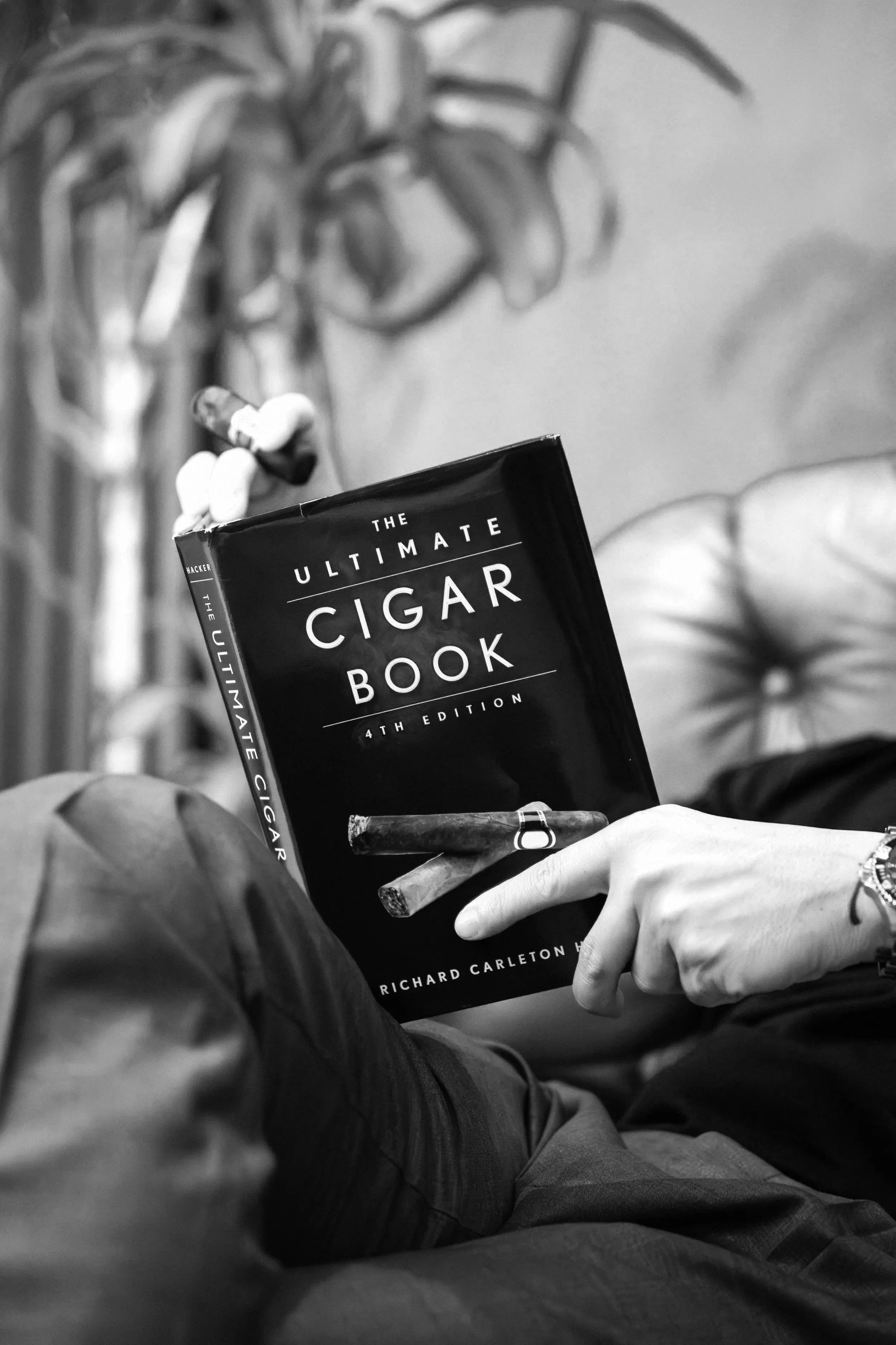 A person sitting down holding a book titled 'The Ultimate Cigar Book' by Richard Carleton Hacker, with a cigar shown on the cover, surrounded by blurred plants in the background.