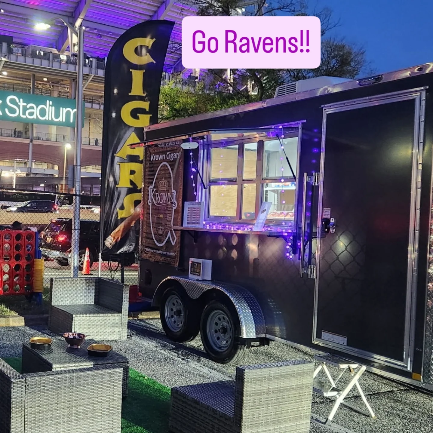 A food truck with a purple sign that says "Go Ravens!!" parked near a stadium at dusk. The truck displays a banner with a cigar and the word "Krown". There are outdoor seating areas with wicker chairs and tables, purple string lights, and a parking lot with cars and an illuminated stadium in the background.