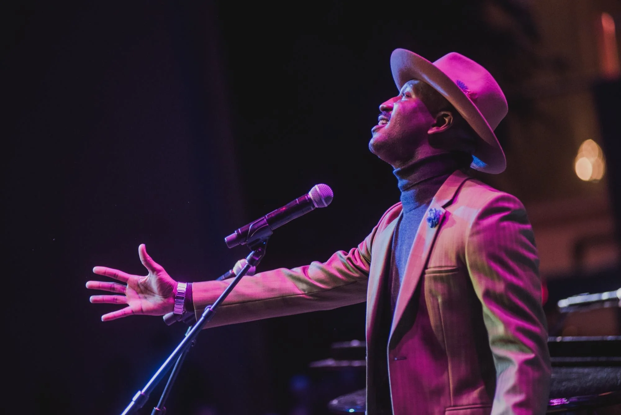 A NewSong Music contestant wearing a blazer, turleneck shirt, and fedora, performs a song onstage.