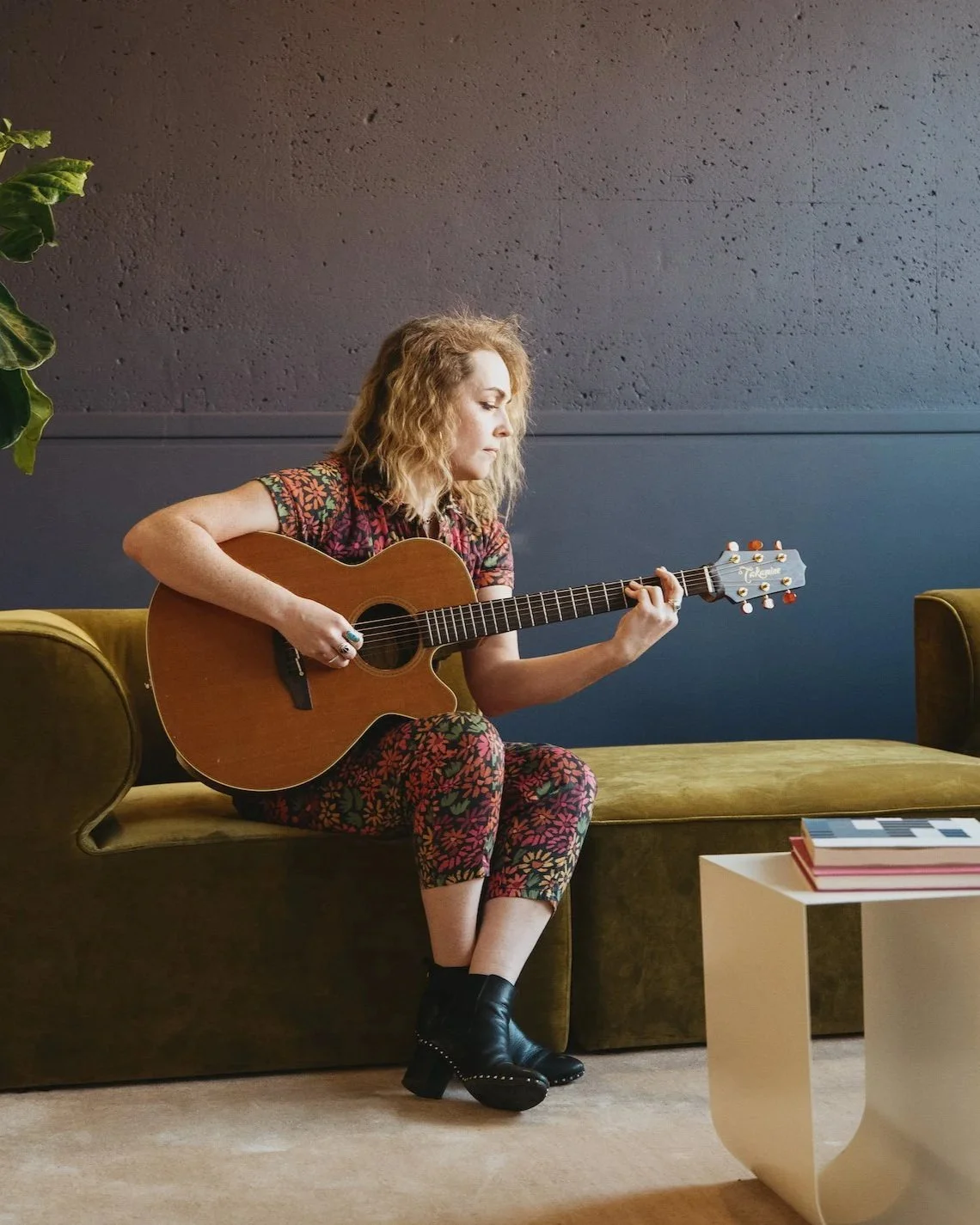 2023 NewSong Music Grand Prize Winner Natalie Price is seated on a green sofa. She wears a floral jumpsuit and holds her guitar.