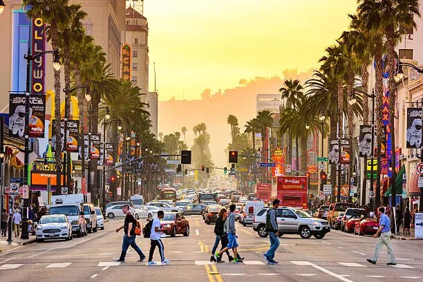 Pedestrians crossing Hollywood Boulevard in Los Angeles at sunset with heavy traffic and city lights in the background.