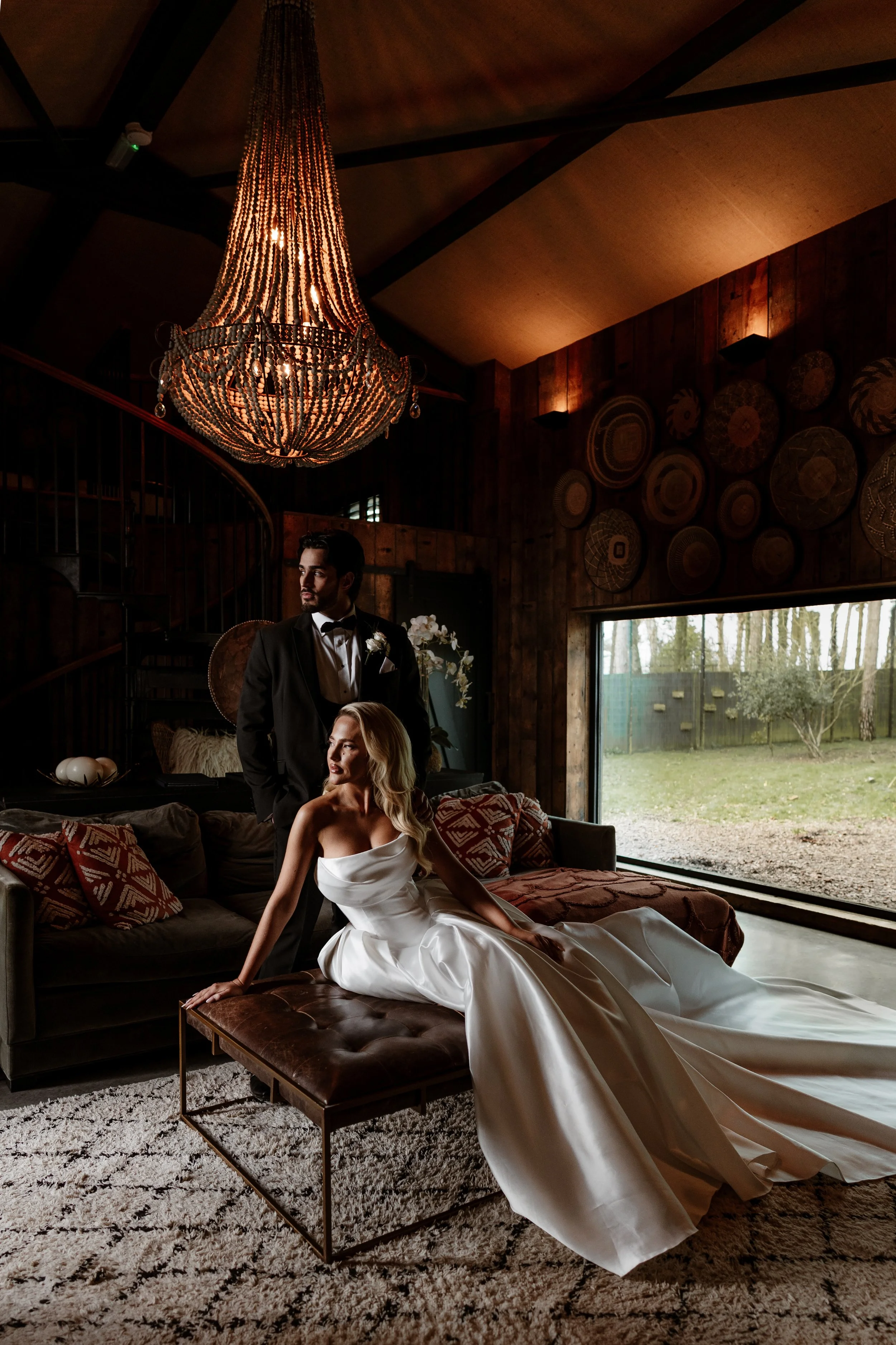 A woman in a white strapless dress sitting on a brown leather ottoman in a living room, with a man in a tuxedo standing behind her, both looking away. The room has dark wood walls, a large chandelier, circular wall art, and a large window showing tre