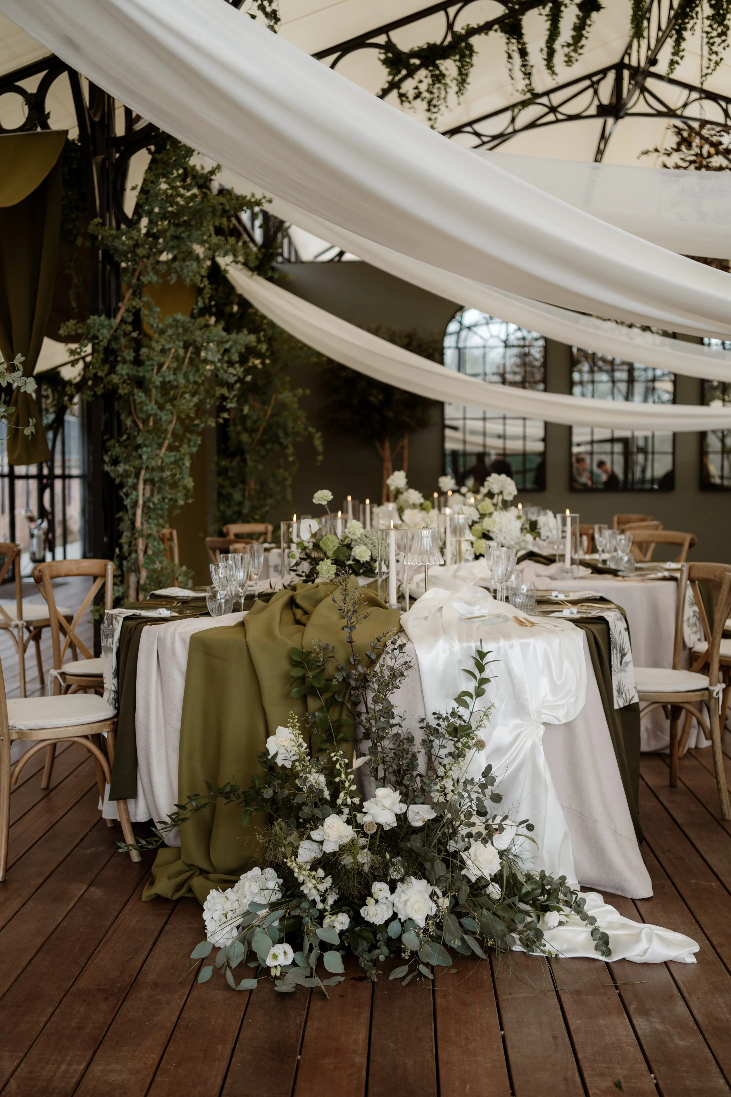 Elegant wedding reception table decorated with white flowers and greenery, topped with draped white and olive-green fabric, in an indoor venue with large windows, black iron structures, and hanging greenery.