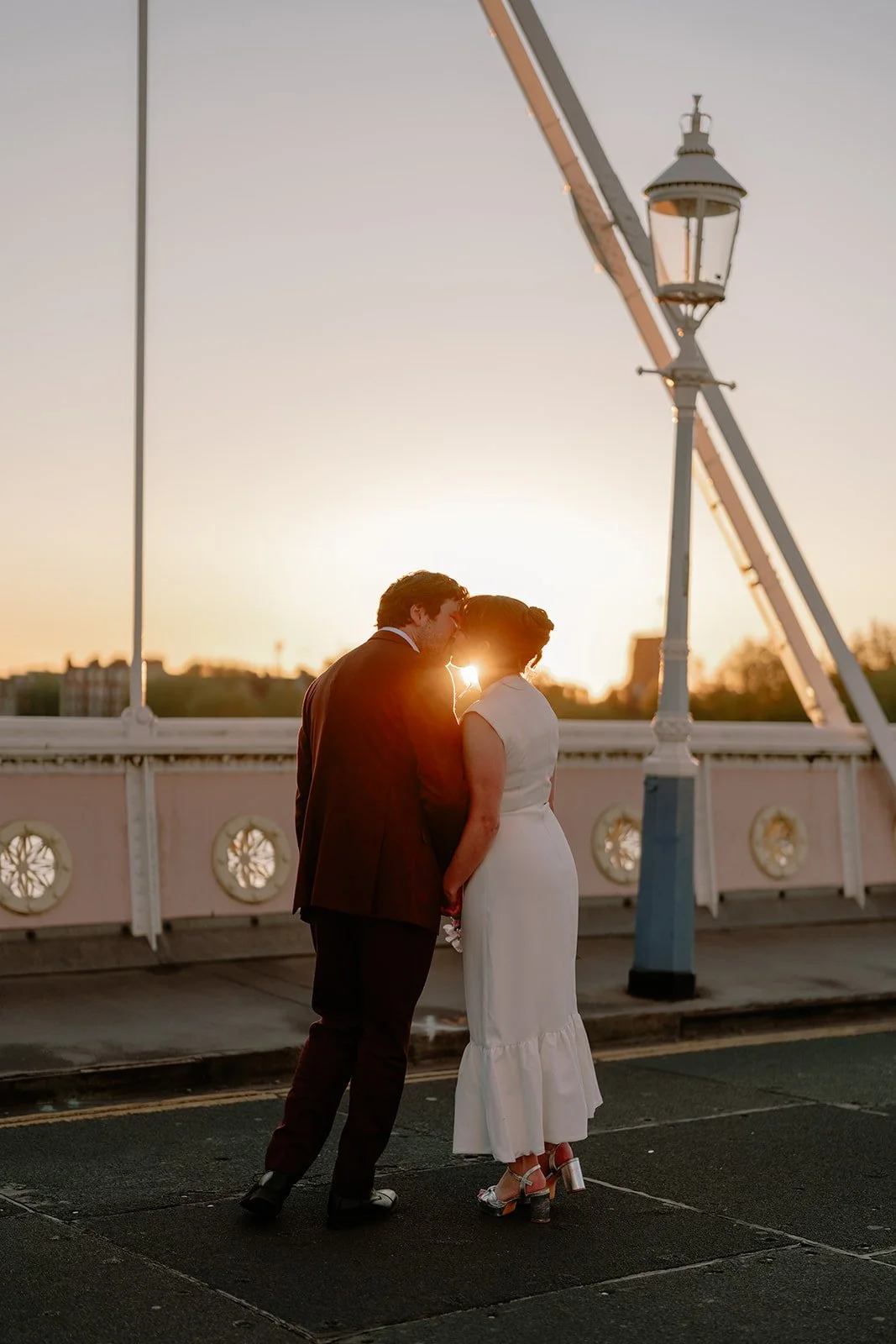 A couple dressed in wedding attire standing close together on a bridge at sunset, holding hands and touching foreheads.