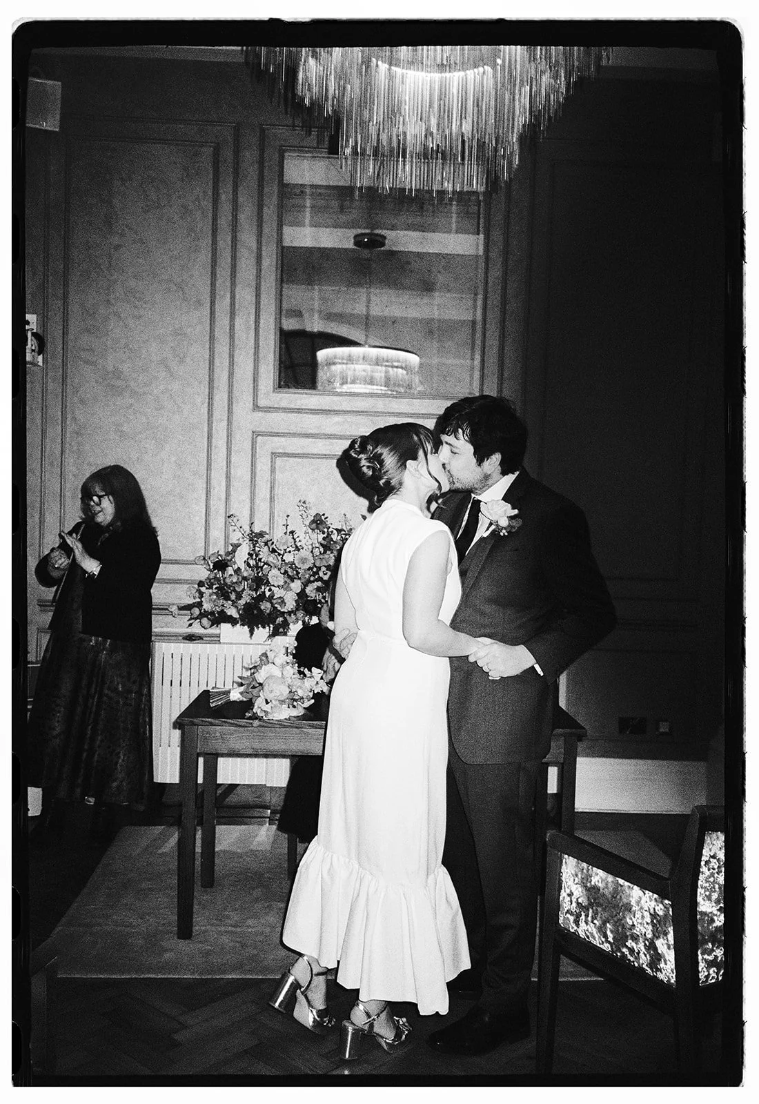 A black and white photo of a couple kissing at their wedding, with a woman in a dress with ruffles and high heels, standing on a wooden floor, behind a table with floral arrangements, in an elegant room with ornate wood paneling and a large mirror.