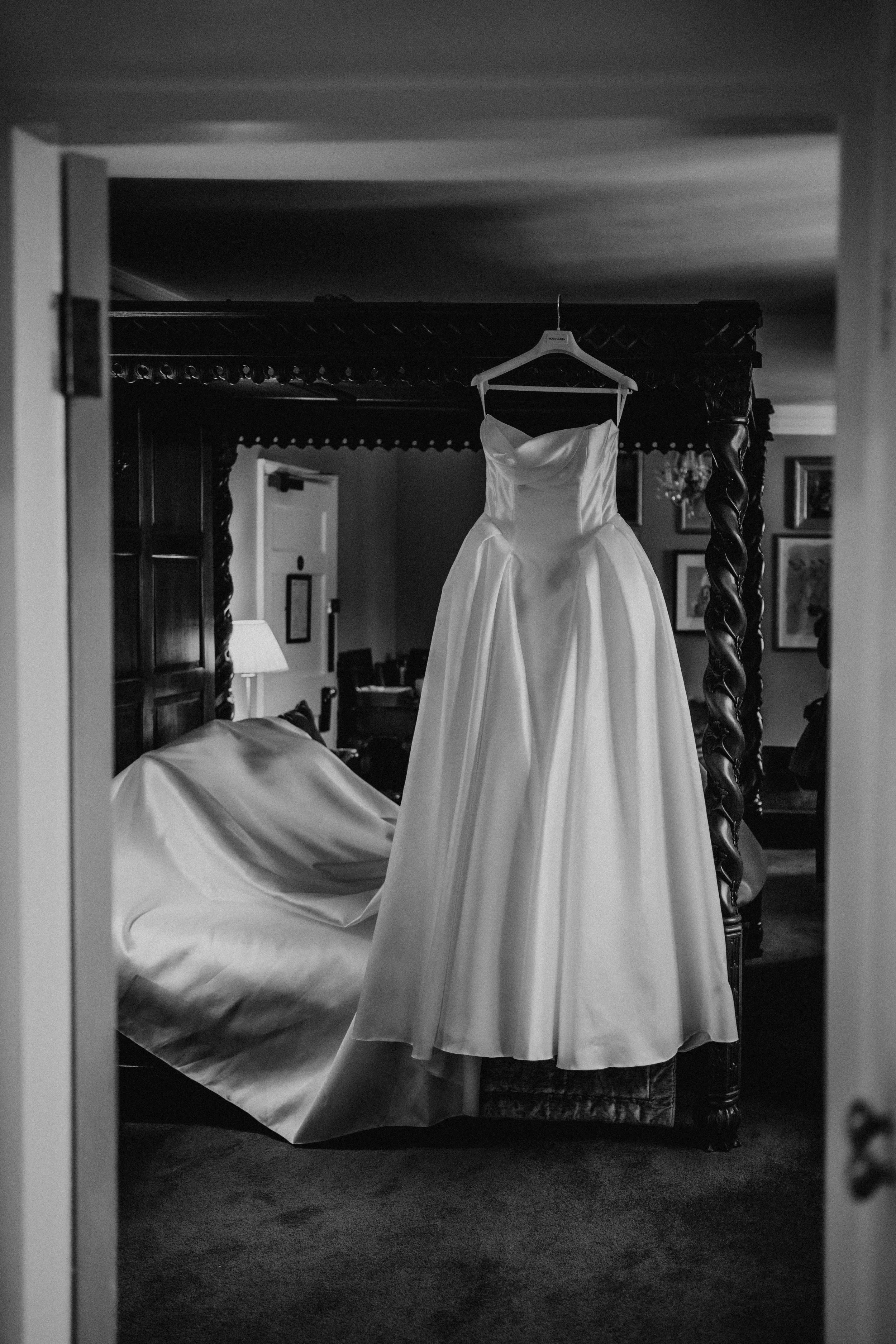 Black and white photo of a wedding dress hanging on a bedpost in a bedroom, viewed through a doorway.