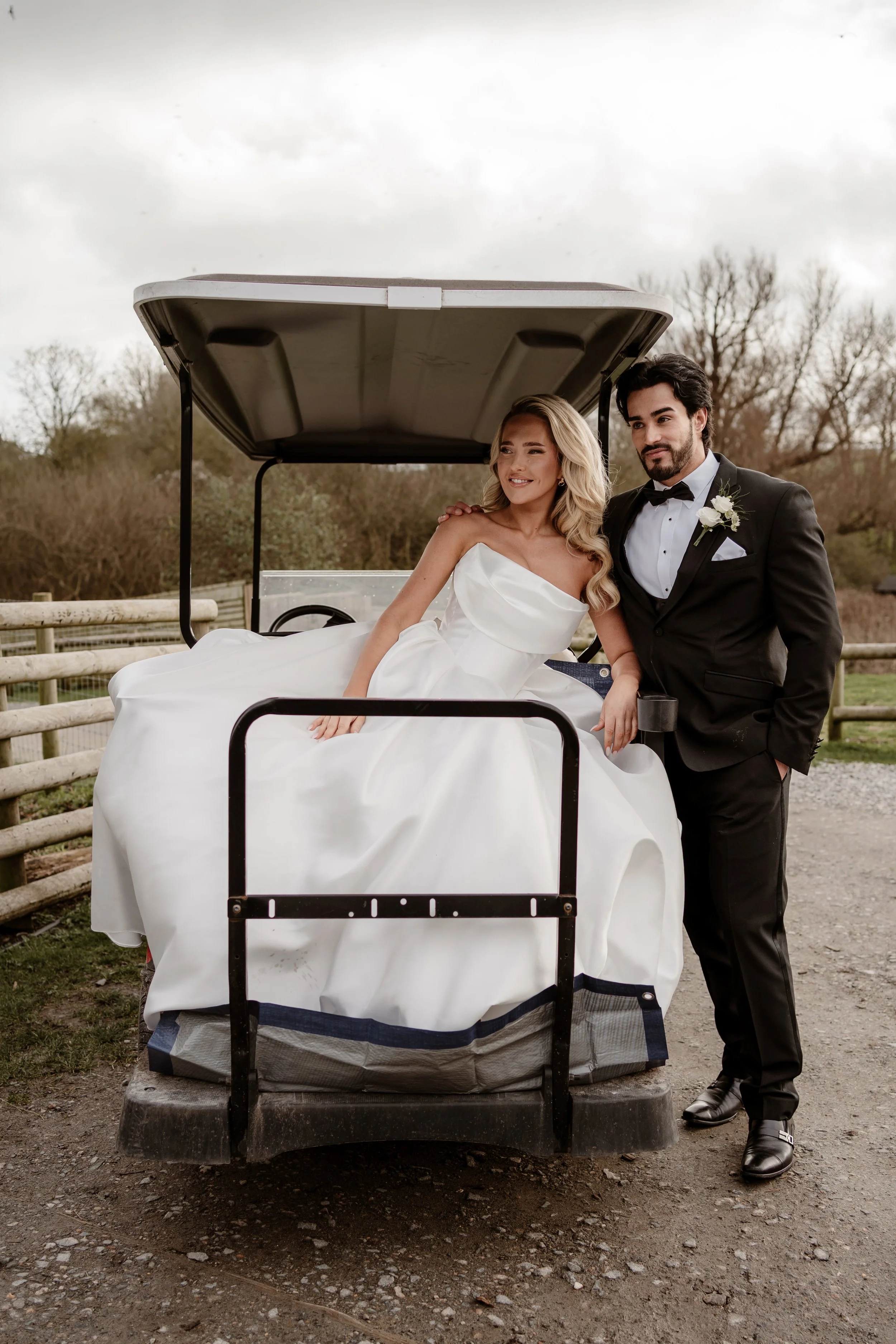 A bride in a white wedding dress and a groom in a black tuxedo with a white boutonniere pose on a golf cart outdoors, with a cloudy sky and bare trees in the background.