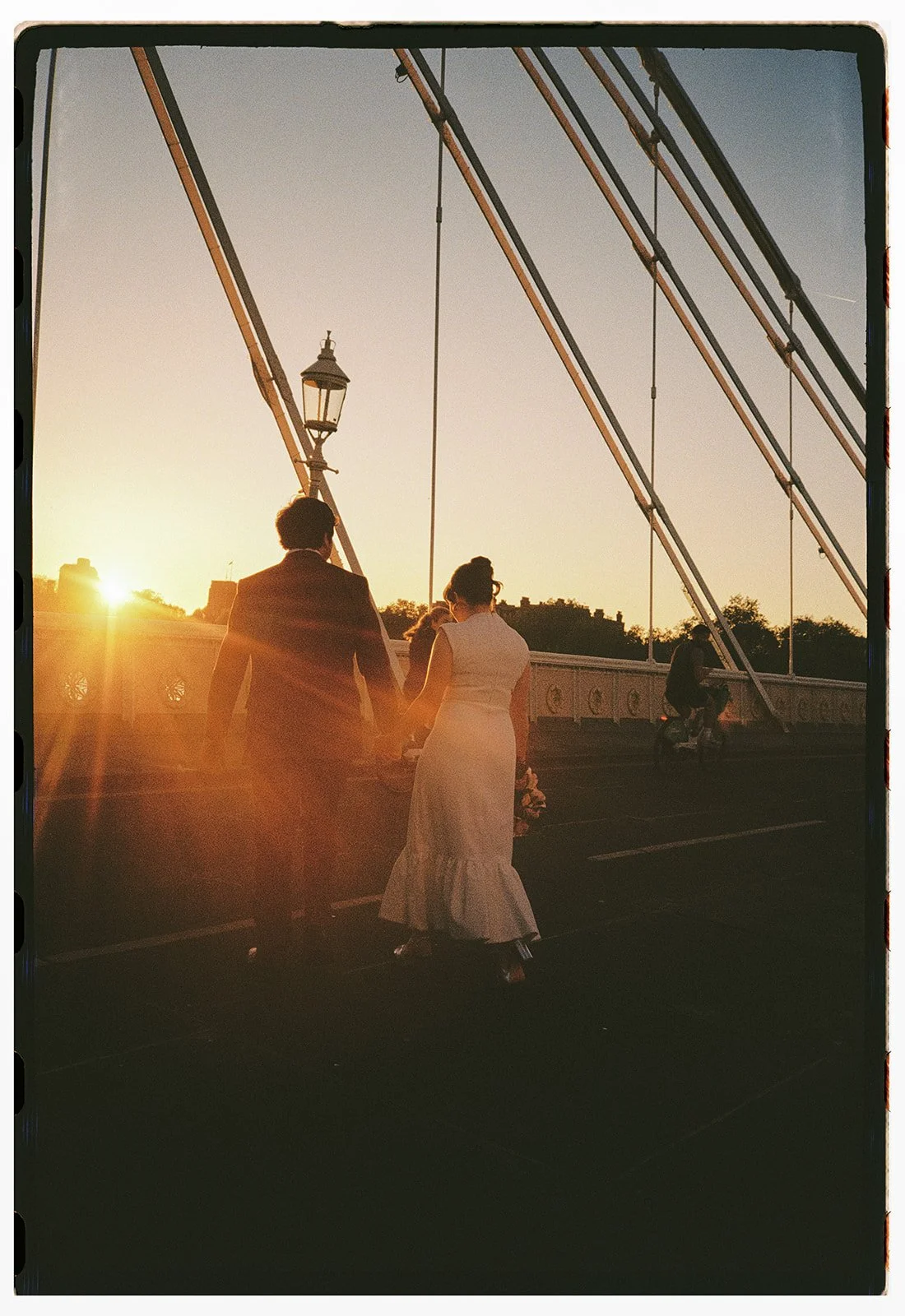 A couple dressed in wedding attire holding hands on a bridge during sunset, with a man on a bicycle in the background.