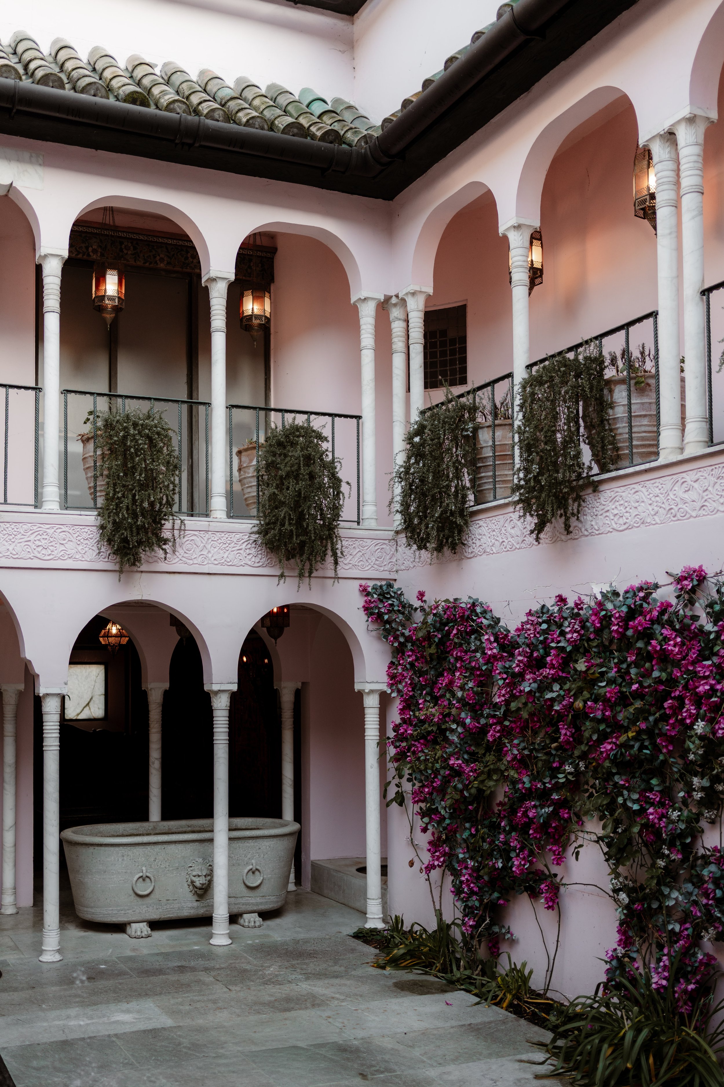 Inner courtyard with pink walls, white columns, hanging lanterns, potted plants, and flowering bushes.