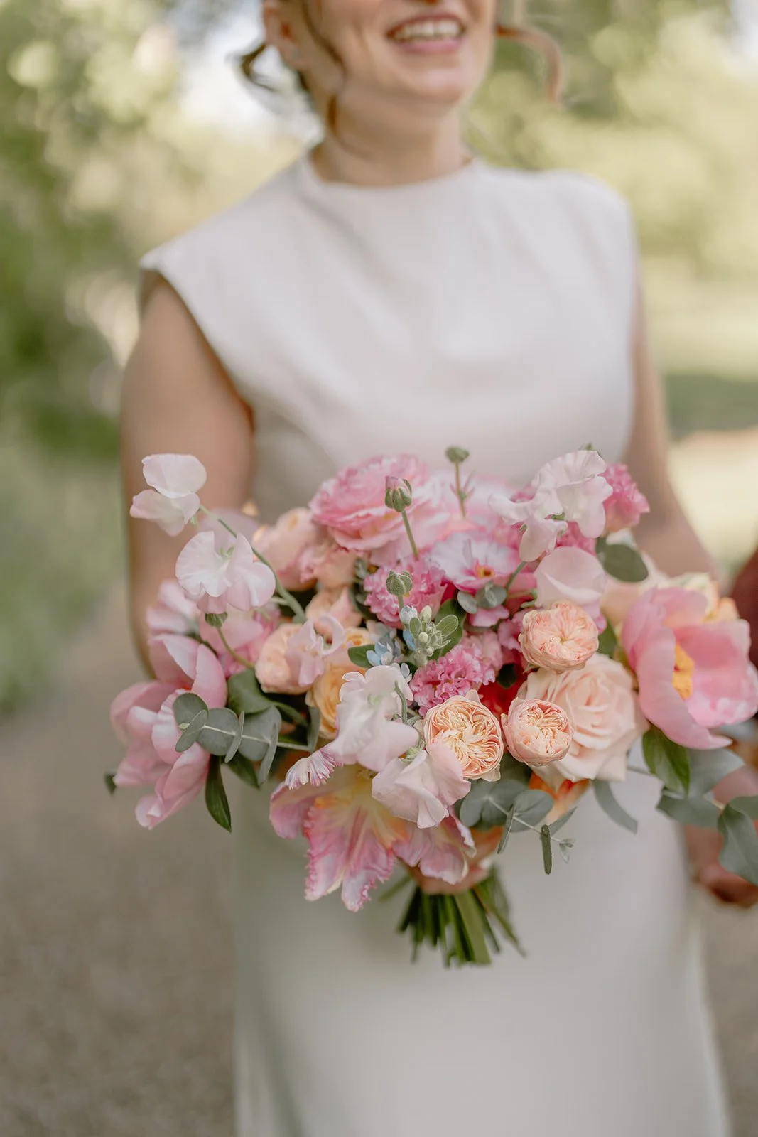 Woman in white dress holding a large bouquet of pink, peach, and white flowers outdoors.