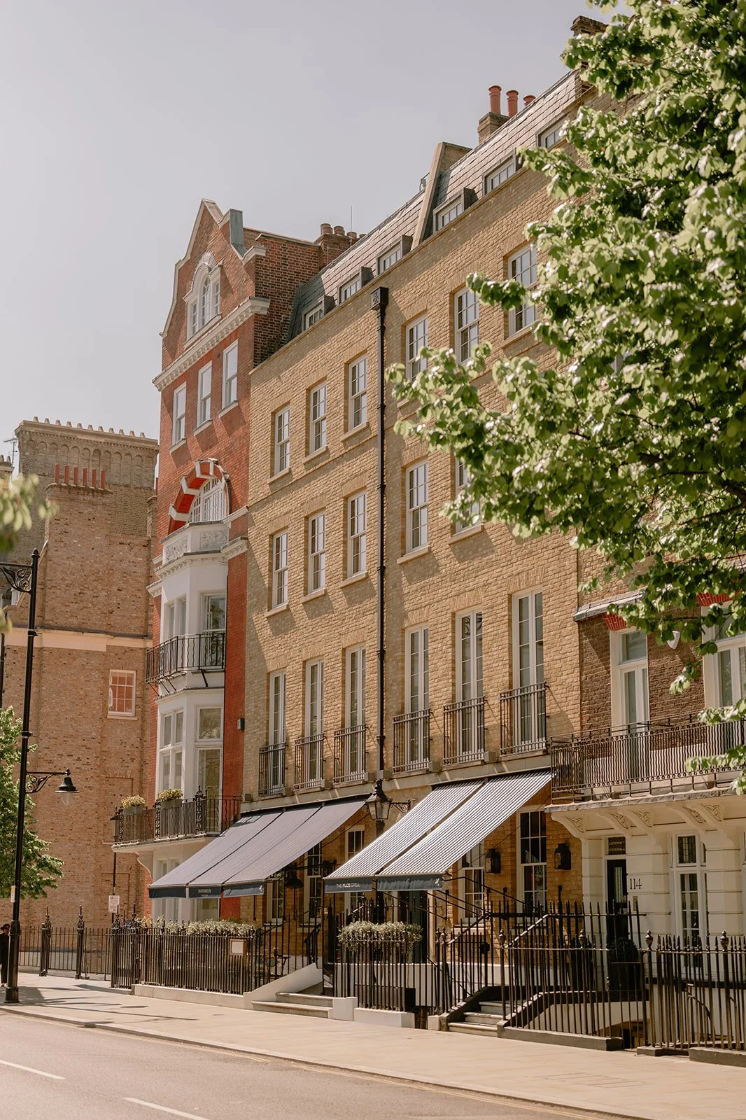 A row of multi-story urban buildings with small balconies and storefronts at street level, featuring awnings and decorative railings, with a sidewalk and trees in the foreground.