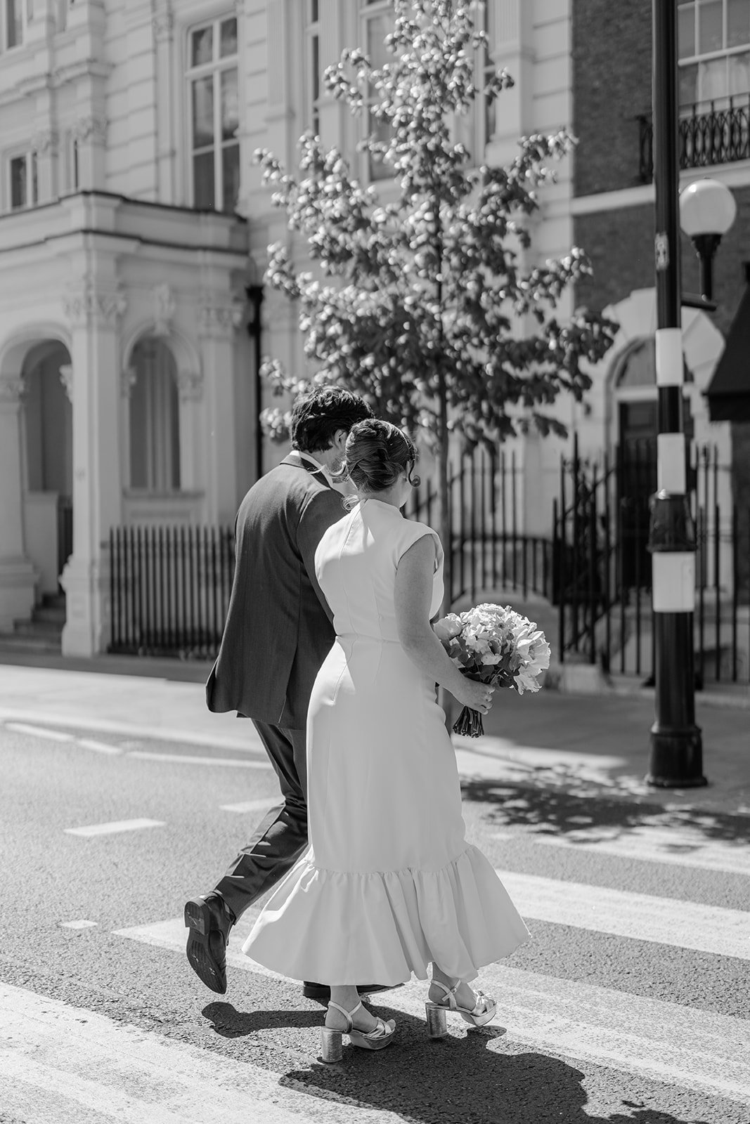 A black-and-white photo of a bride and groom walking on a city street, with the bride holding a bouquet of flowers and wearing high-heeled sandals, and the groom focused on her. There are residential buildings, a tree, and street lamps in the backgro