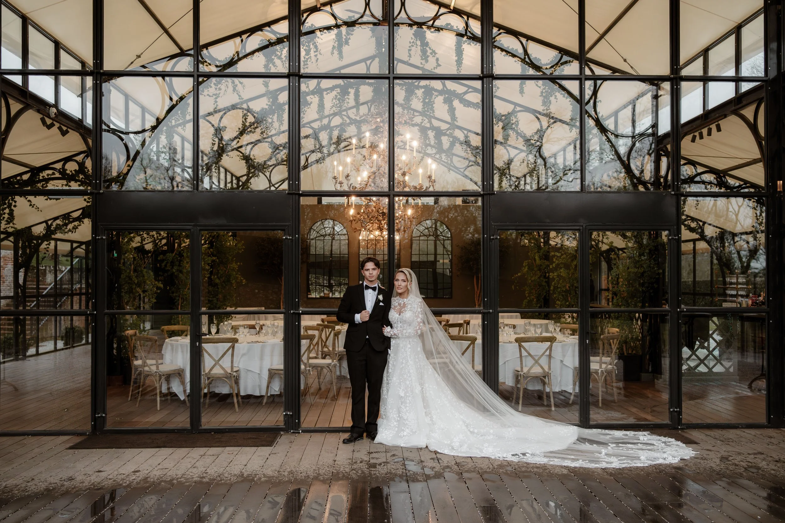 A bride and groom standing inside a glass-walled greenhouse or conservatory during their wedding, with the groom in a tuxedo and the bride in a lace wedding gown and veil.