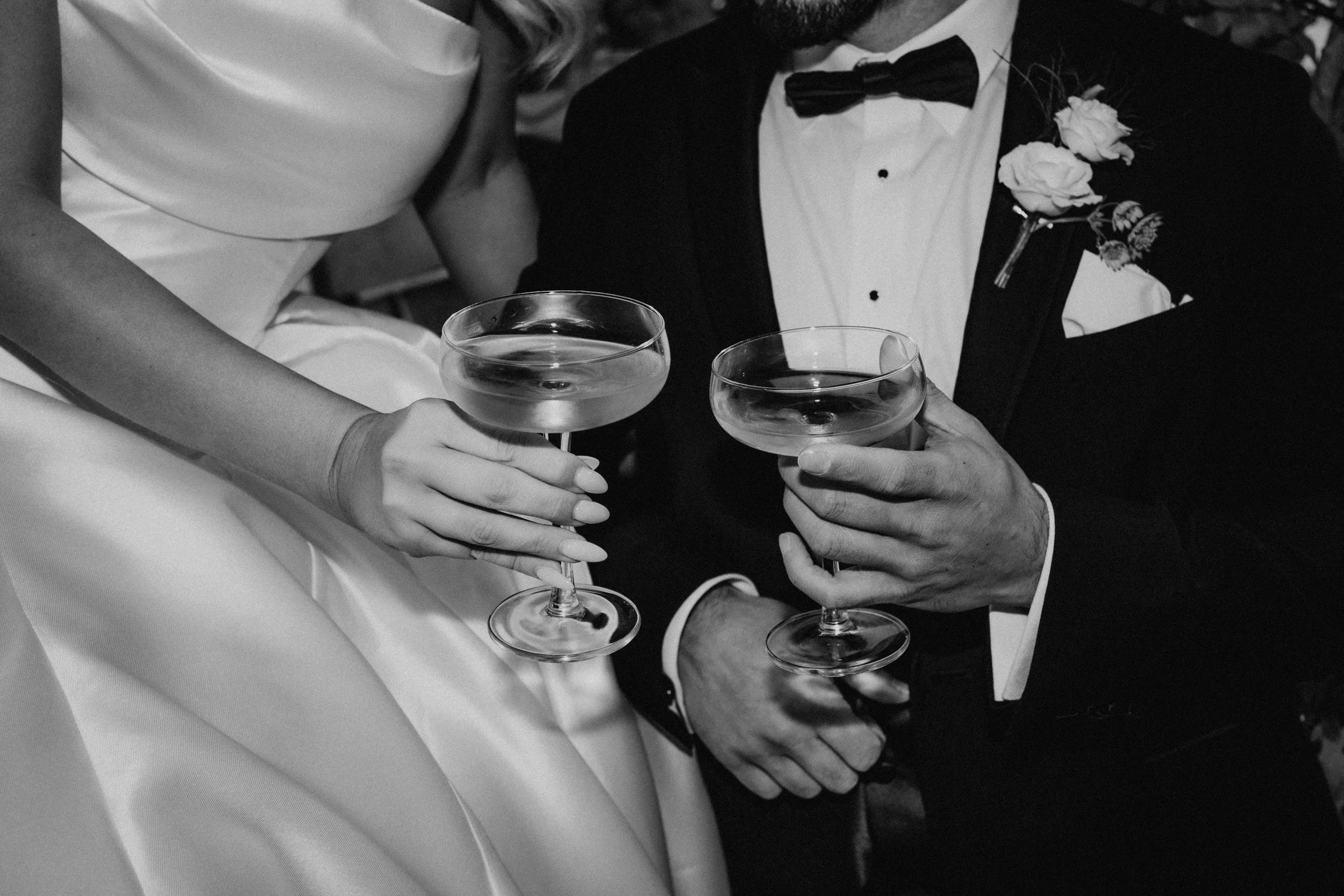 Black and white photo of a couple dressed in wedding attire holding champagne glasses, with hands touching, celebrating an occasion.