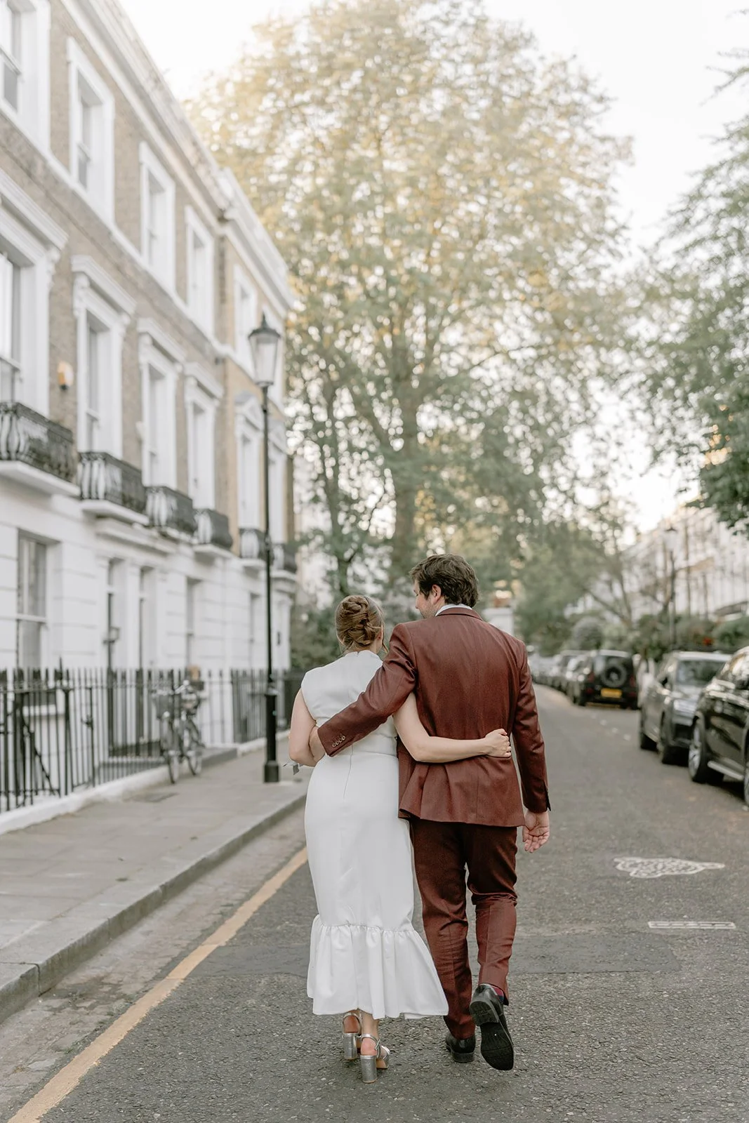 A man and woman walking arm-in-arm down a city street. The woman is in a white dress and high heels, and the man is in a brown suit. Trees and parked cars line both sides of the street.