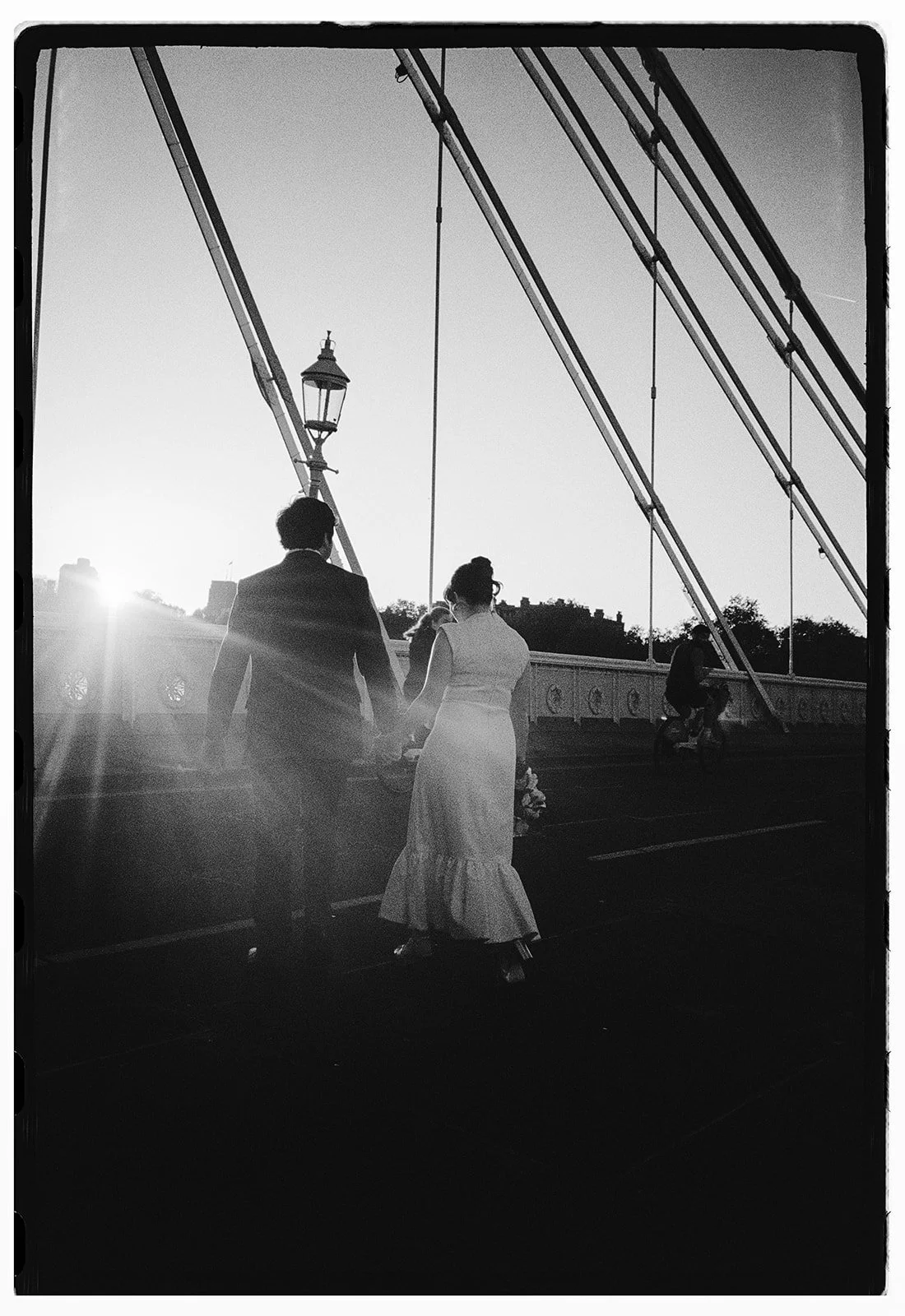 A black-and-white photo of a couple holding hands on a bridge at sunset, with a cyclist passing by and a vintage street lamp.