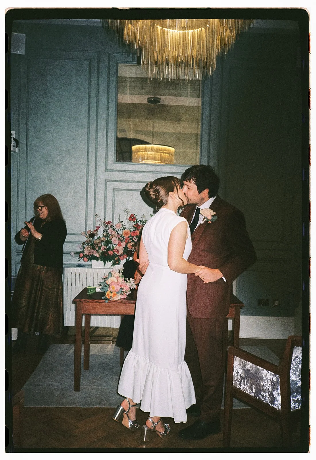 A wedding couple sharing a kiss in a decorated room with flowers and a chandelier, with a woman in the background looking at her phone.