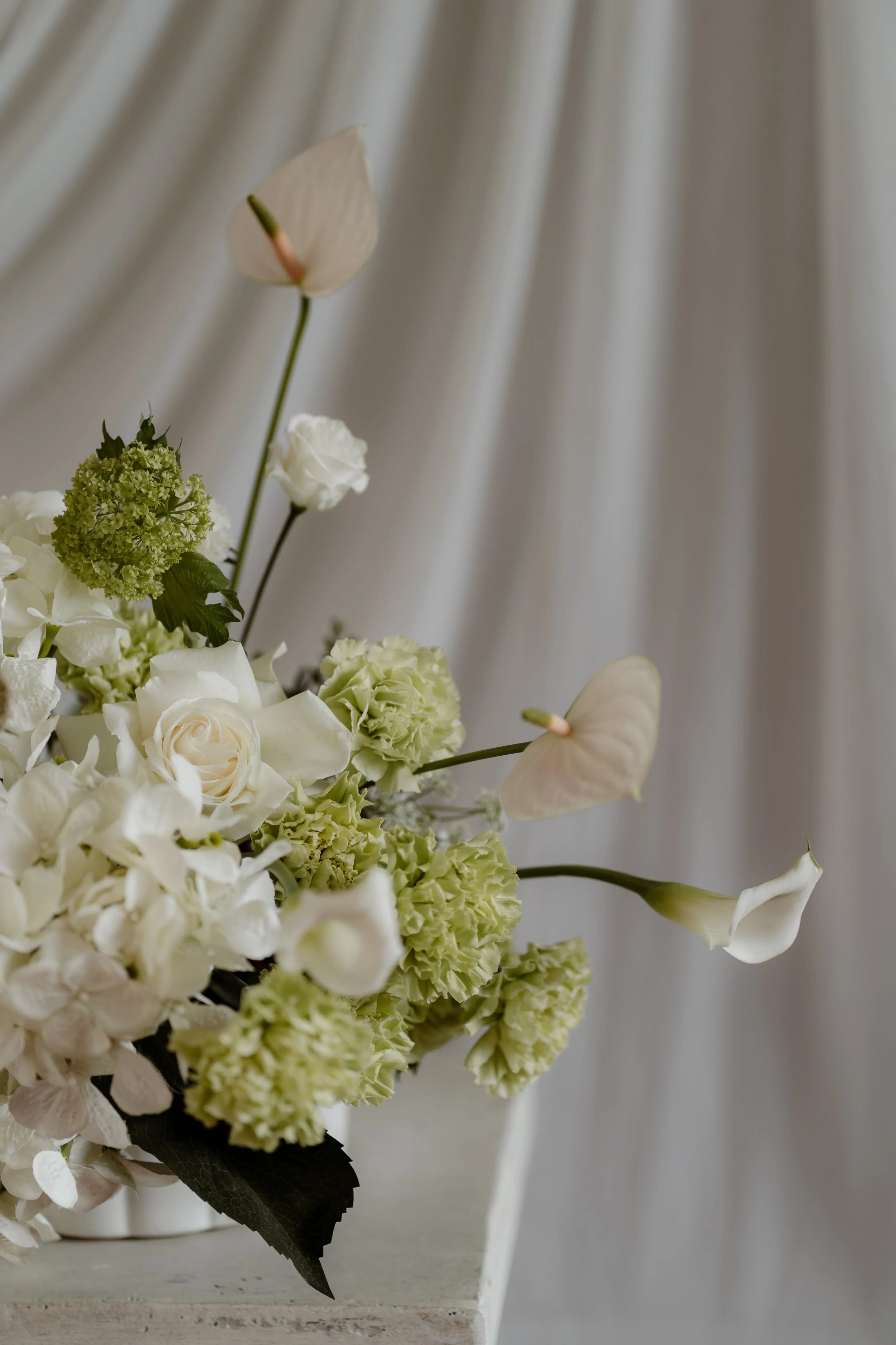 A bouquet of white and pale green flowers, including roses, hydrangeas, and calla lilies, with a white butterfly resting on a flower, against a soft, light fabric background.