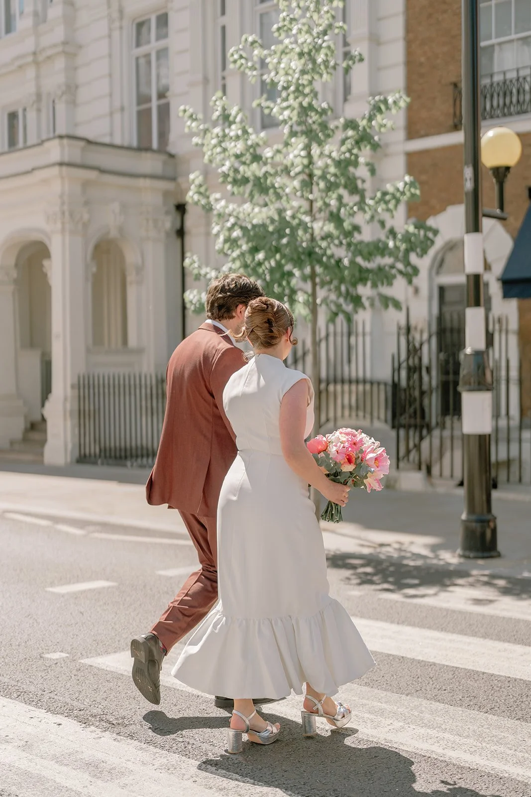 A couple dressed in wedding attire crossing a street, with the woman holding a bouquet of pink and white flowers. They are in an urban setting with white and brick buildings in the background and a tree with white flowers nearby.
